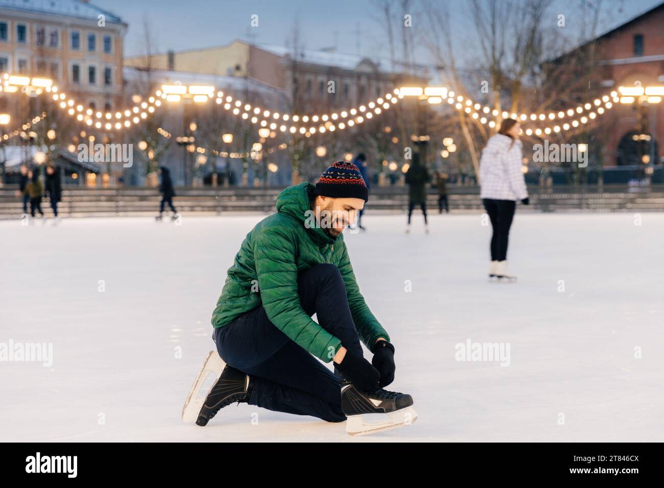 Homme attachant ses patins à glace sur une patinoire éclairée festive au crépuscule, avec des patineurs derrière. Banque D'Images