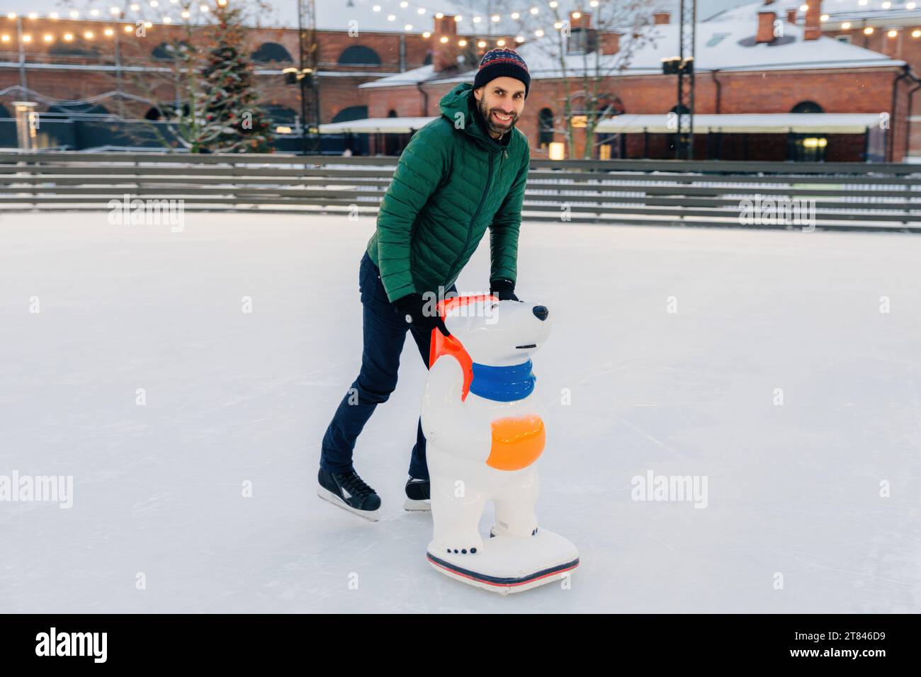Homme heureux patinant avec une aide en forme d'ours sur une patinoire extérieure au crépuscule Banque D'Images