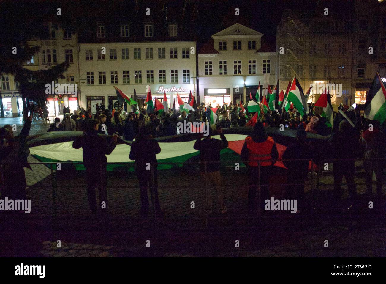 Flensburg, Schleswig-Holstein, Pro-Palästina-démonstration à Flensburg. Gesamtbild Demonstranten halten große Fahne von Palästina, die weiteren Personen bilden Halbkreis, in der Mitte nicht zu erkennen in gehüllte Laken weiße Puppen mit roter Farbe beschmiert, die tote Personen symbolisieren sollen. Aufnahme vom 18.11.2023, Flensburg, Holm/Südermarkt *** Flensburg, Schleswig Holstein, manifestation pro Palestine à Flensburg vue générale manifestants tenant un grand drapeau palestinien, les autres personnes forment un demi-cercle, au milieu des poupées méconnaissables enveloppées dans des draps blancs enduits de r Banque D'Images