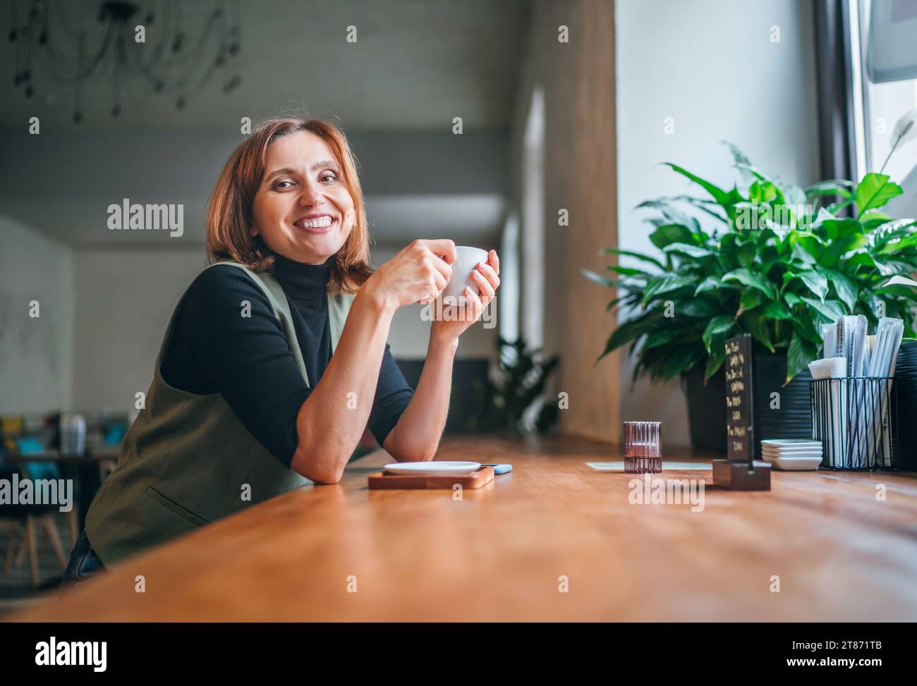 Portrait de femme caucasienne brune sincèrement souriante assise à table dans un café confortable avec une petite tasse de café aromatique, jonchée de lumière du matin de fenêtre Banque D'Images
