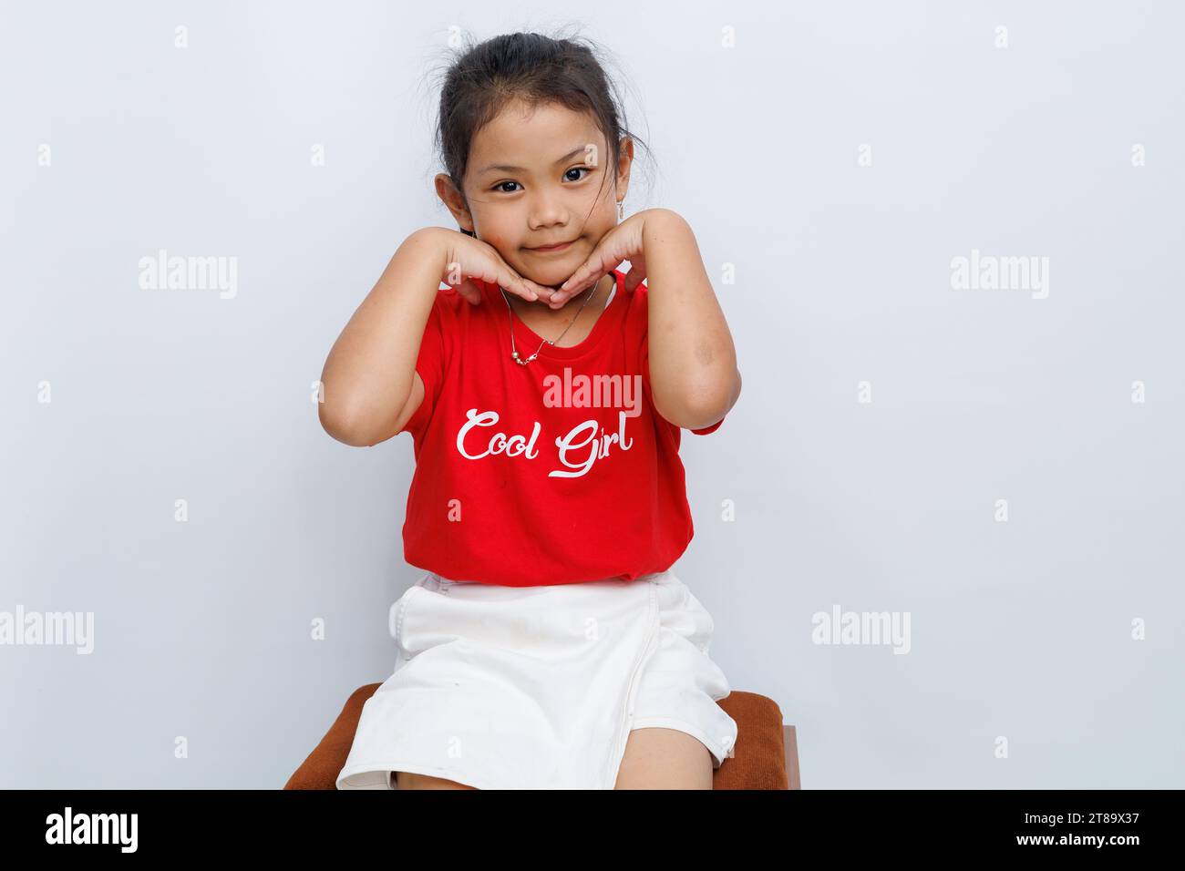 Petite fille asiatique assise devant un fond blanc et les paumes extérieures de ses mains soutenant ses joues. en souriant en regardant la caméra. Banque D'Images