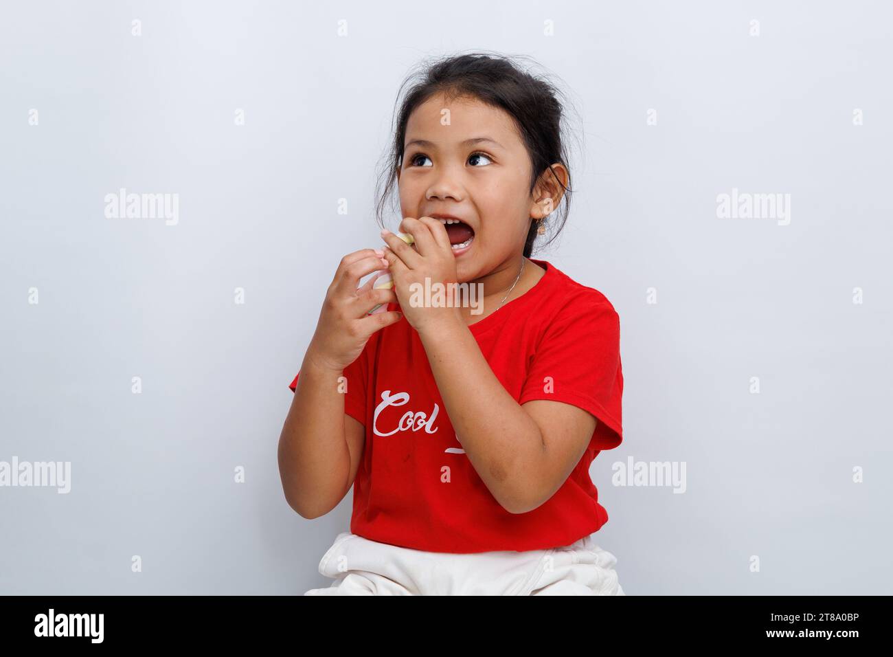 Une petite fille asiatique portant un t-shirt rouge mord une fraise avec une expression de curiosité sur le goût de la fraise. Banque D'Images