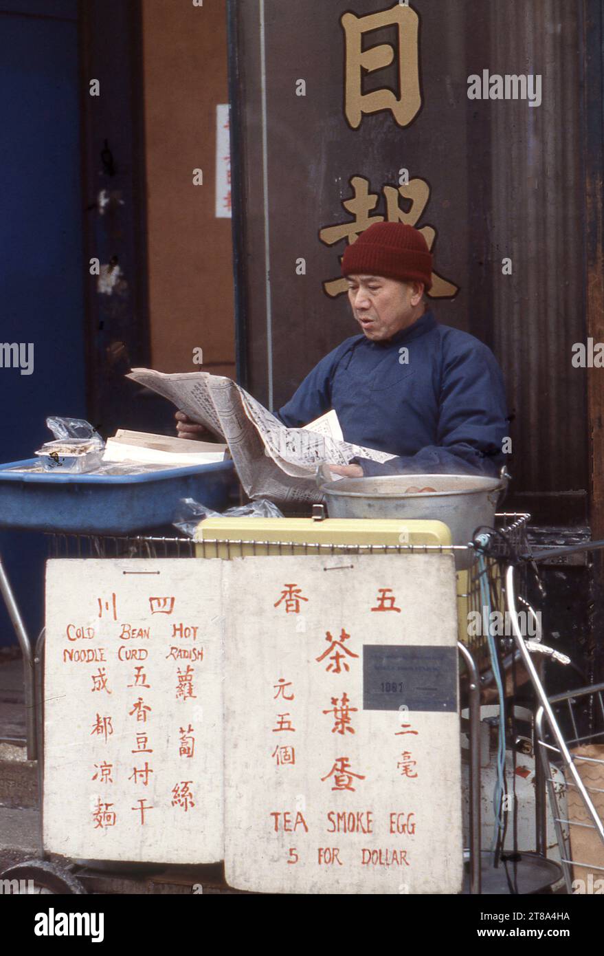 Un sino-américain lit un journal derrière son stand où il vend des œufs fumés au thé, des nouilles froides, du caillé de haricots et des radis chauds. Sur Canal Street dans le quartier chinois de New York en janvier 1979. Banque D'Images