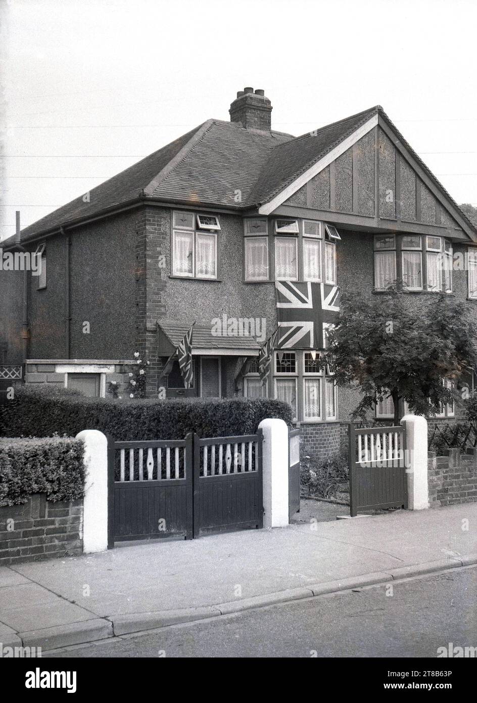 1953, historique, vue de face d'une maison de banlieue semi-détachée construite dans les années 1930, rendu de galets, avec un grand drapeau de l'Union Jack à l'extérieur, sous la fenêtre, avec des drapeaux plus petits sur le porche, Angleterre, Royaume-Uni, en célébration du couronnement de la Reine. Banque D'Images