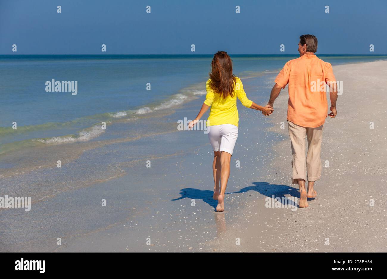 Vue arrière d'un couple heureux homme et femme courant, tenant les mains sur une plage déserte avec un ciel bleu clair lumineux Banque D'Images