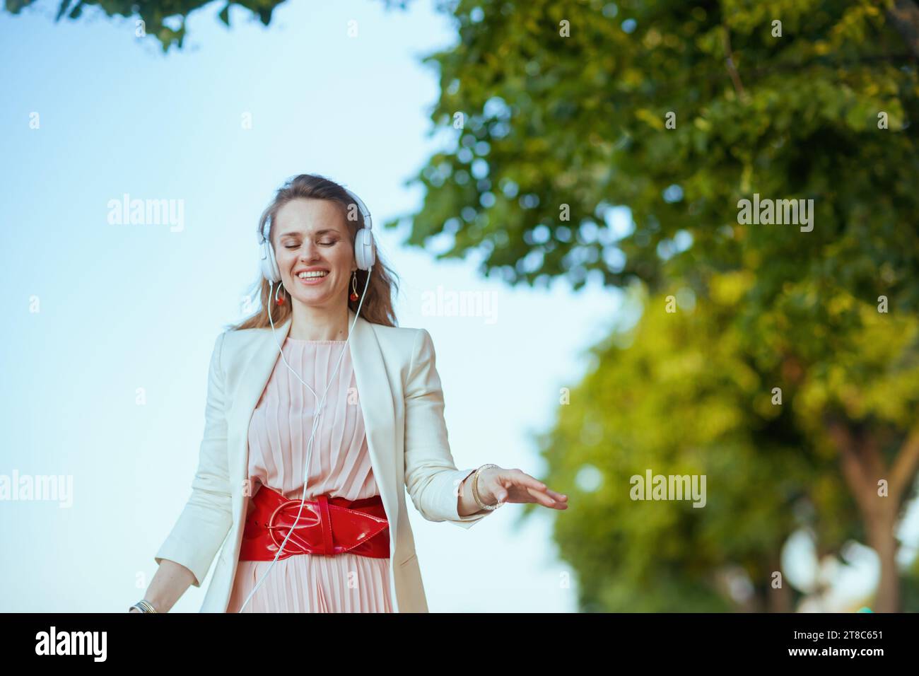 souriant femme tendance de 40 ans en robe rose et veste blanche dans la ville écoutant la musique avec des écouteurs. Banque D'Images