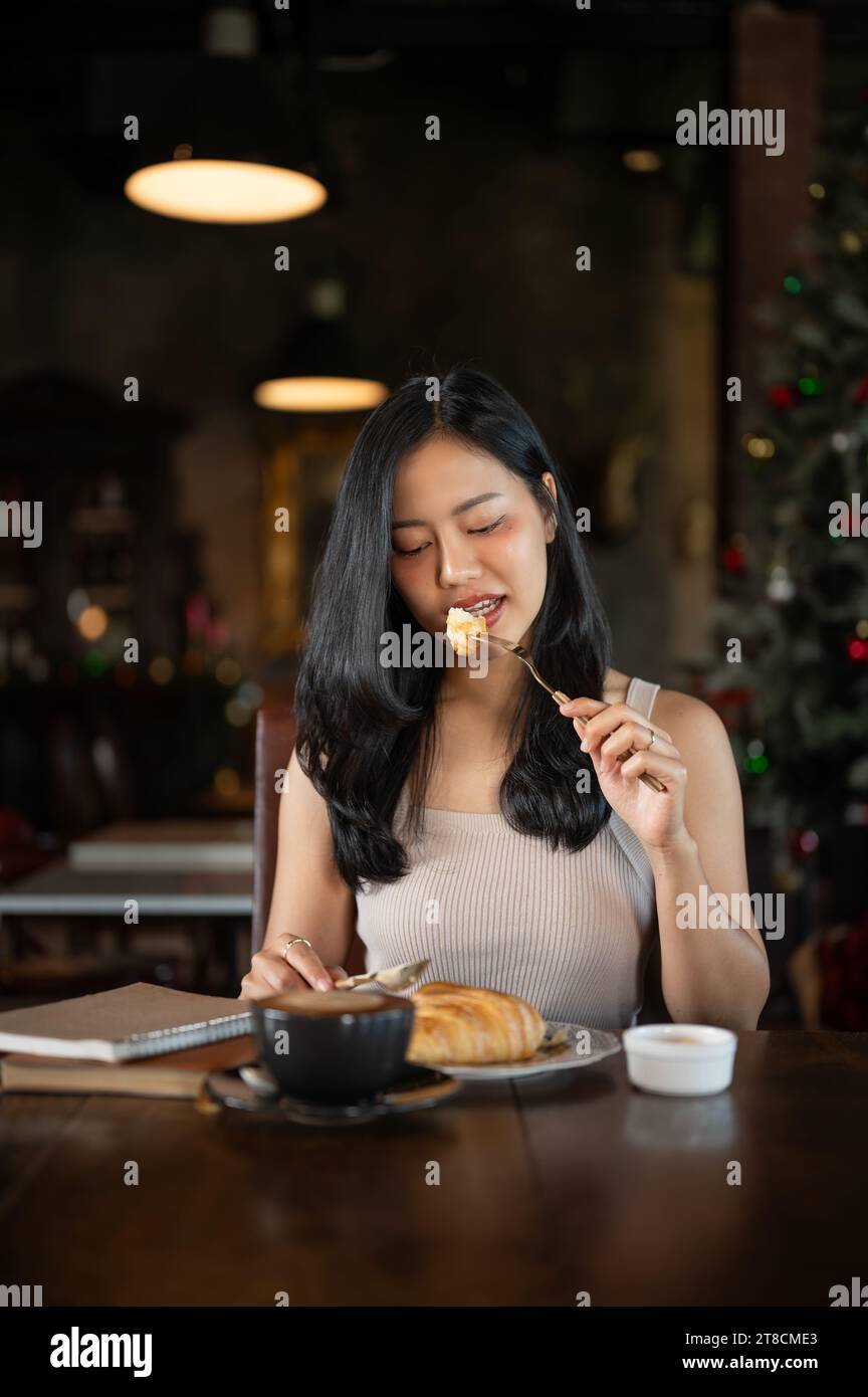Élégante jeune femme asiatique mangeant un croissant pour le petit déjeuner avec latte chaud dans un salon de luxe sombre. Petit déjeuner du matin avec café. Banque D'Images
