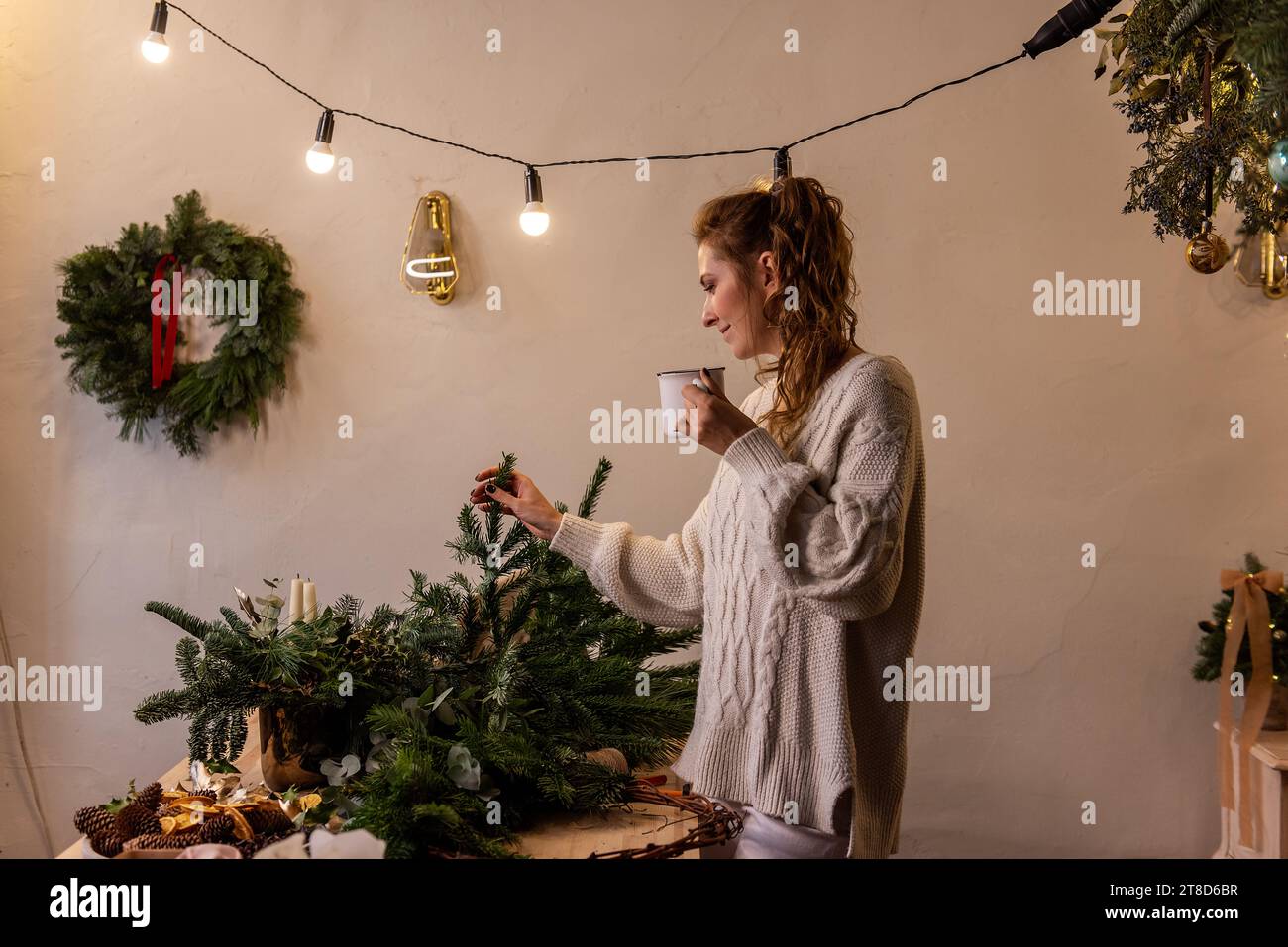 Jeune femme fleuriste atelier fait la couronne de Noël, boit le thé chaud de tasse blanche pendant la pause entre le travail. Petite entreprise pendant les vacances. Hiver A. Banque D'Images