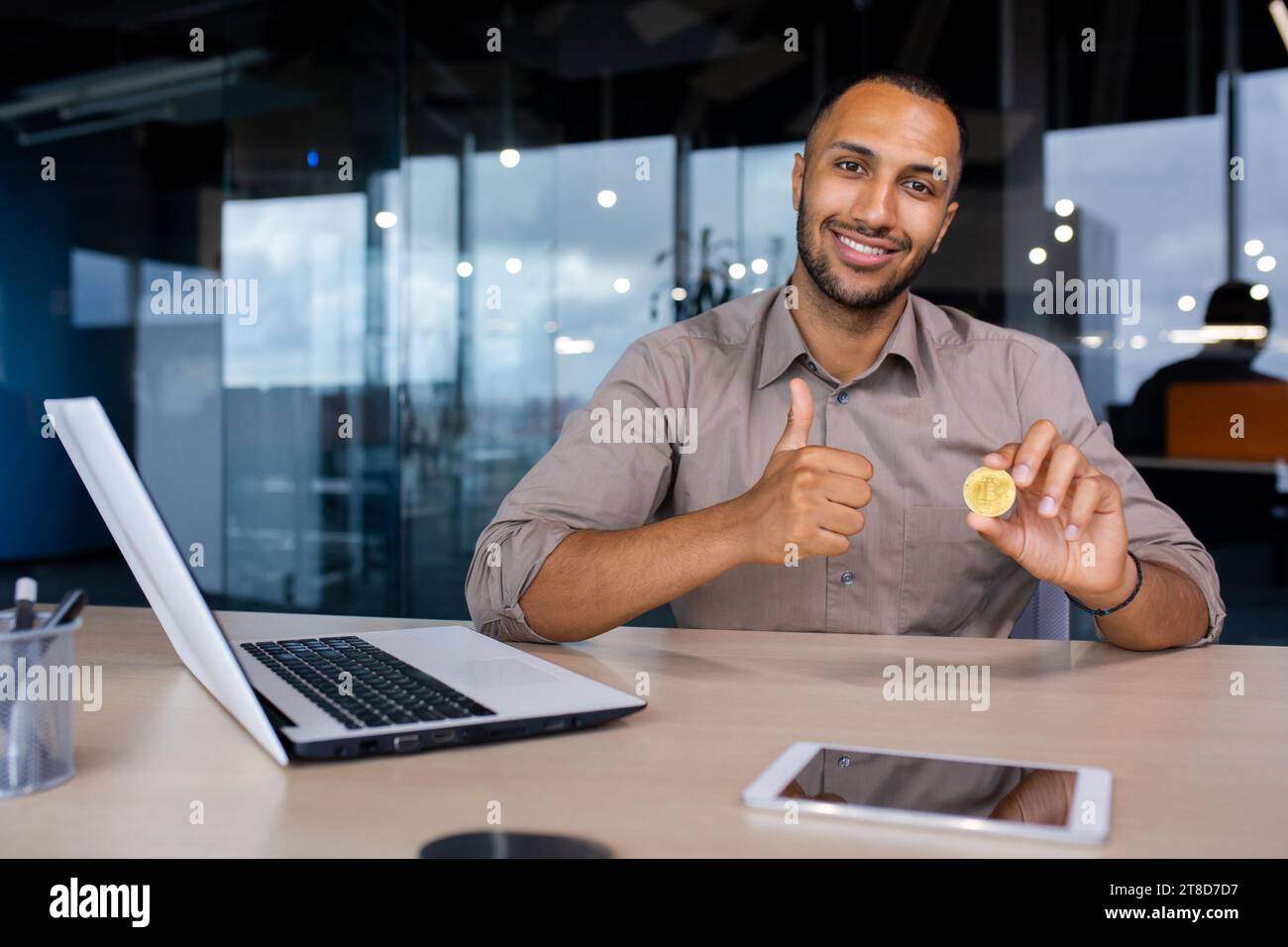 Heureux homme d'affaires afro-américain assis dans le bureau tenant le bitcoin virtuel de crypto-monnaie montrant comme pouce vers le haut. Banque D'Images