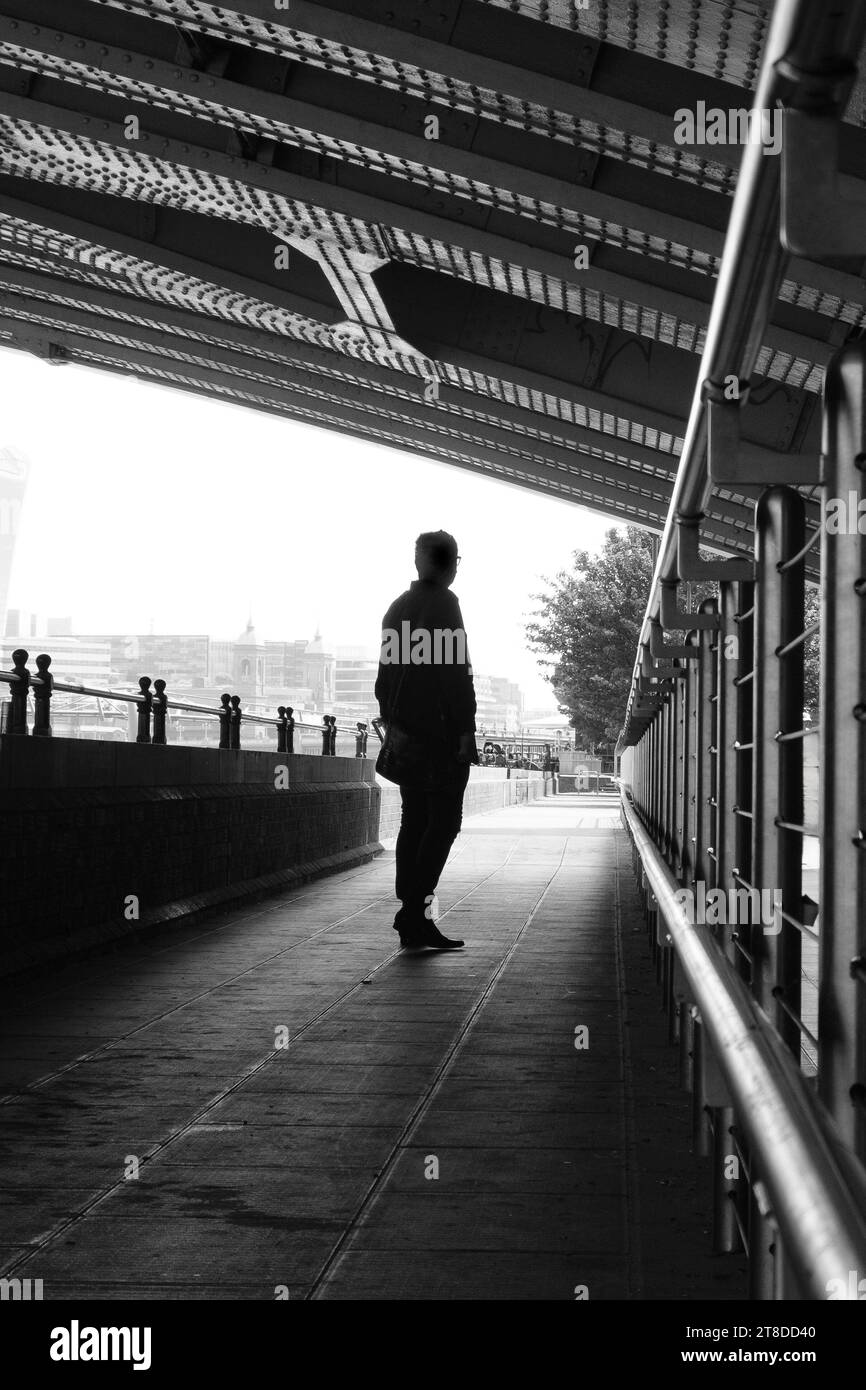 Image en noir et blanc de la silhouette d'un homme debout dans un passage urbain avec toit bas, lignes directrices et point de fuite contre le ciel lumineux. Banque D'Images