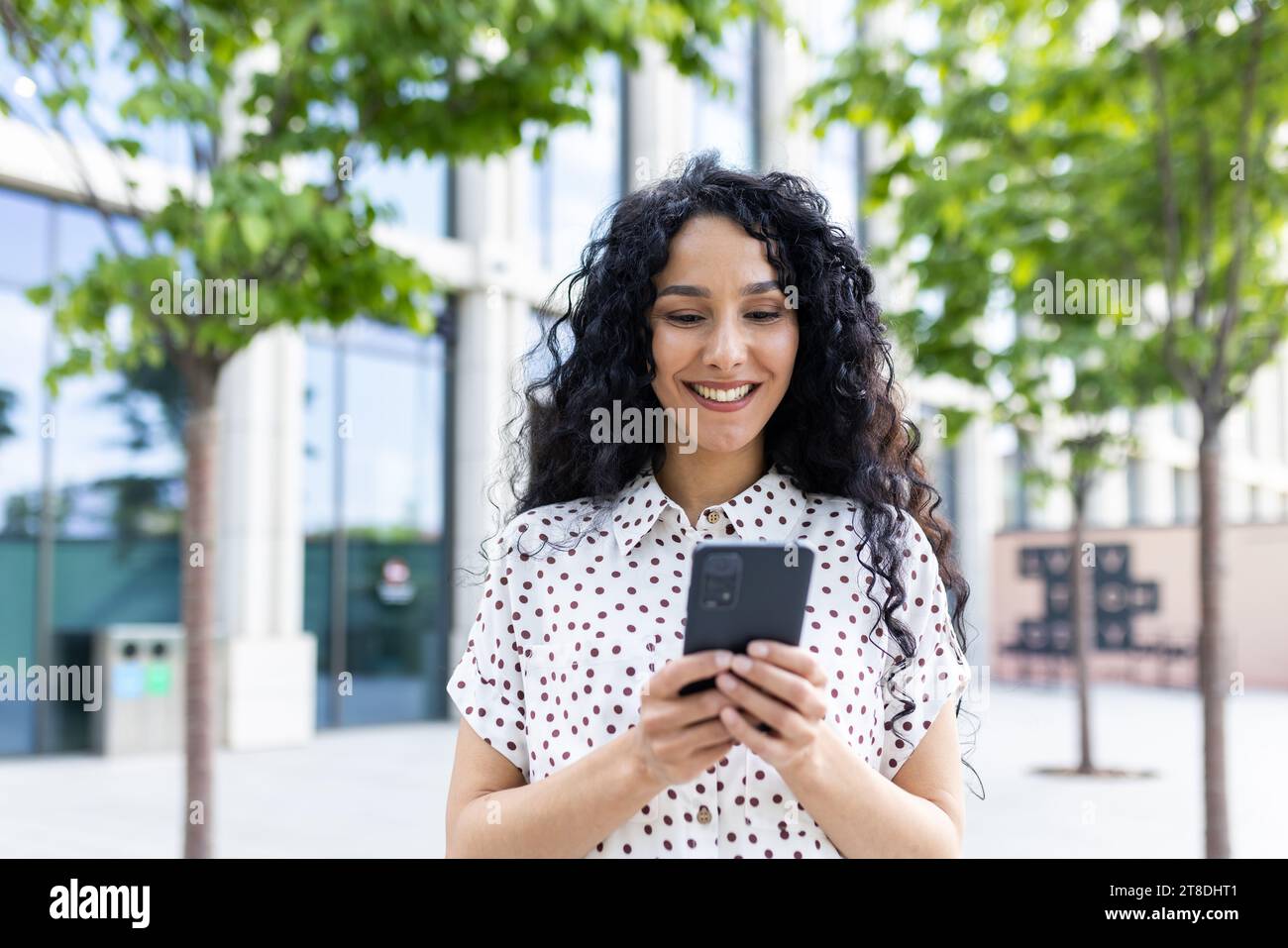 Une jeune belle femme se promène dans la ville avec un téléphone dans les mains, sourit contentement, utilise une application pour smartphone, navigue sur des pages Internet, tape un message texte, les réseaux sociaux. Banque D'Images