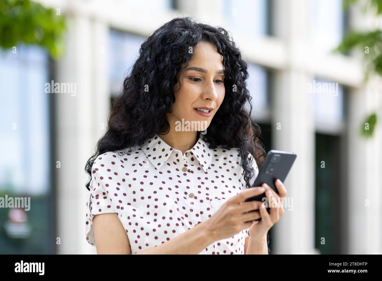 Une jeune belle femme se promène dans la ville avec un téléphone dans les mains, sourit contentement, utilise une application pour smartphone, navigue sur des pages Internet, tape un message texte, les réseaux sociaux. Banque D'Images