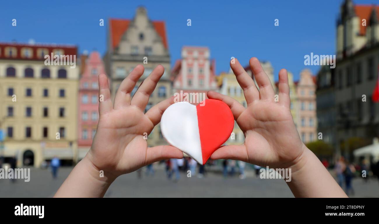 Un cœur rouge-blanc aux couleurs du drapeau national de la Pologne dans les mains d'un enfant sur fond de la place de la vieille ville. Jour de l'indépendance Banque D'Images
