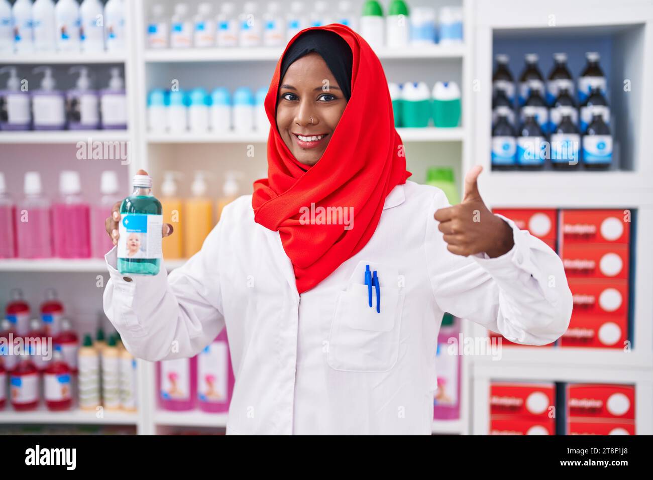 Jeune femme arabe travaillant à la pharmacie pharmacie tenant sirop souriant heureux et positif, pouce vers le haut faisant excellent et signe d'approbation Banque D'Images