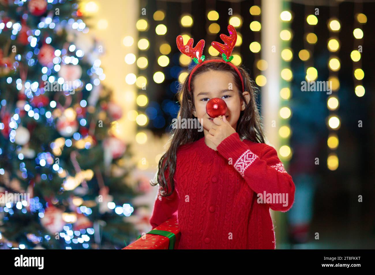 Enfant avec nez de renne et bois la veille de Noël. Petite fille tenant une boule rouge et un cadeau de Noël. Cadeaux de Noël ouverts pour les enfants. Vacances d'hiver fu Banque D'Images