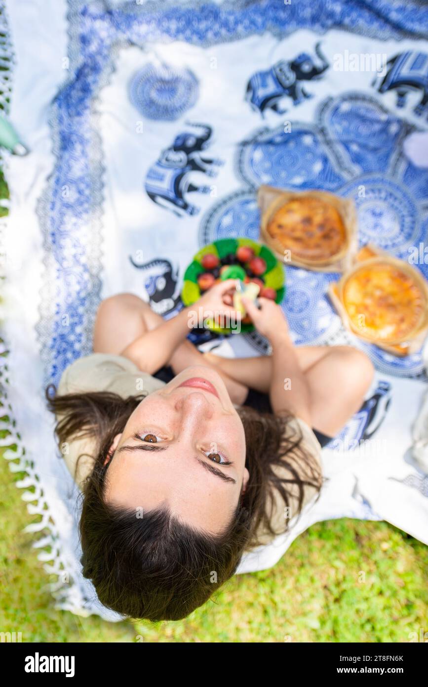 Une femme profite d'un pique-nique seule, allongée sur le dos sur une couverture, tenant des fruits dans ses mains. Banque D'Images