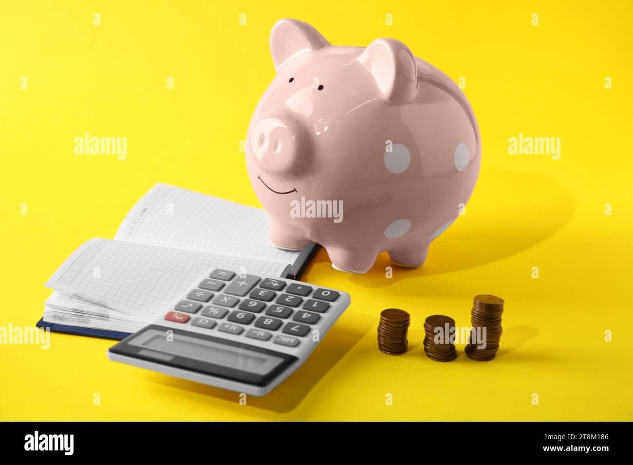 Calculatrice, tirelire rose, cahier et pièces empilées sur fond jaune Banque D'Images