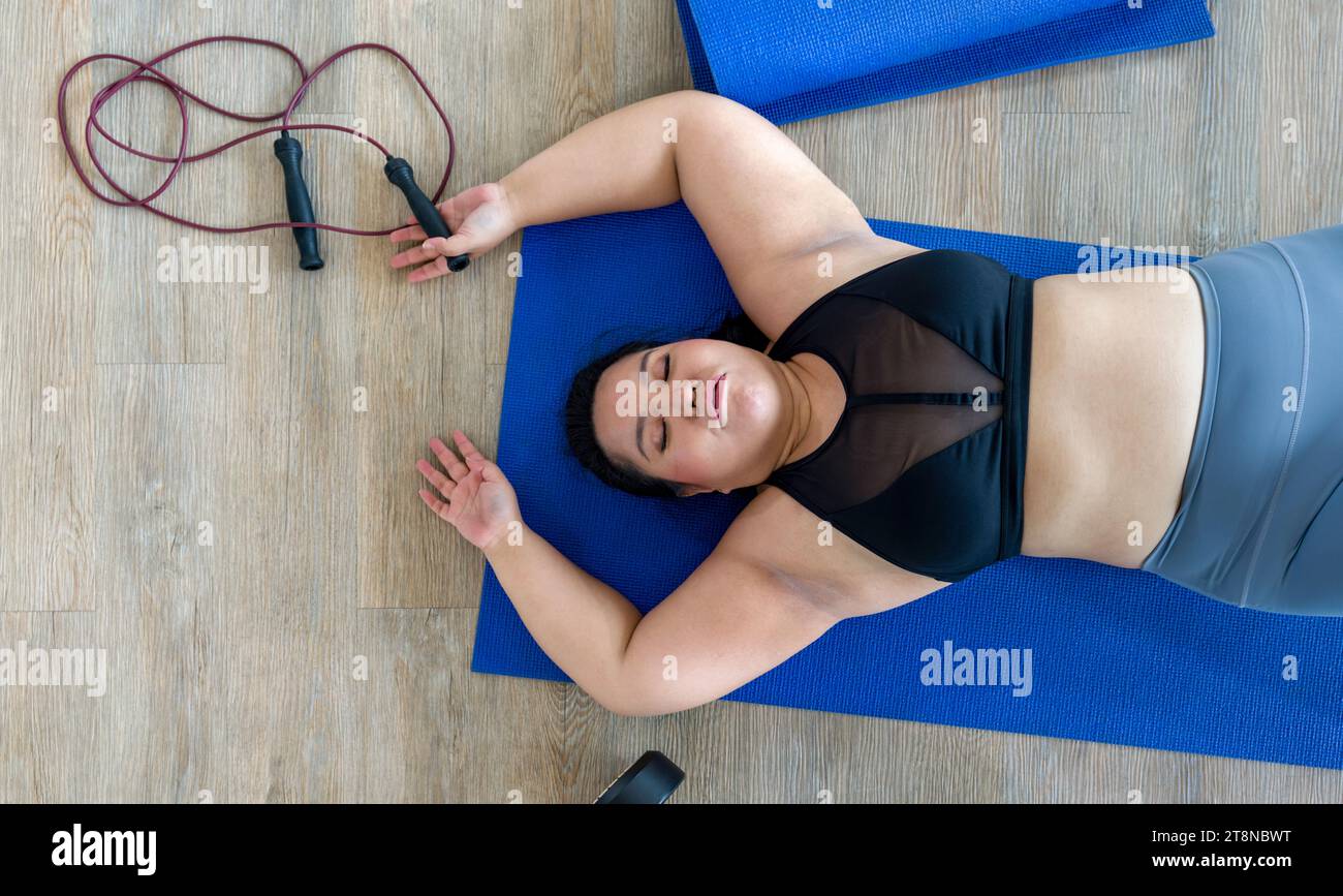 Une femme grinçante semble fatiguée, couchée sur un tapis bleu vif, ses bras reposant à côté d'haltères et de cordes à sauter. Banque D'Images
