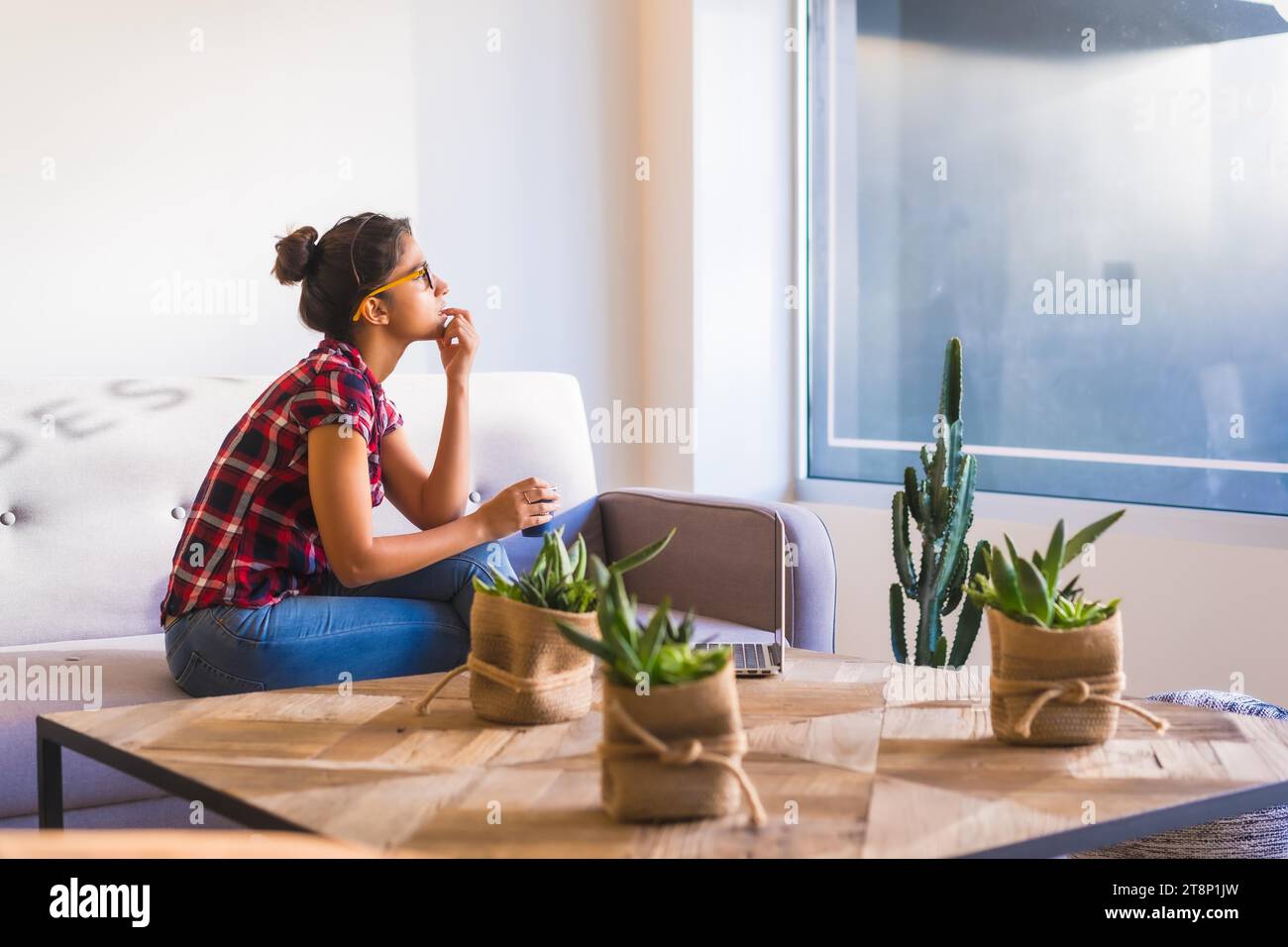 Femme rêveuse regardant par la fenêtre à la maison tout en utilisant un ordinateur portable et boire du café Banque D'Images