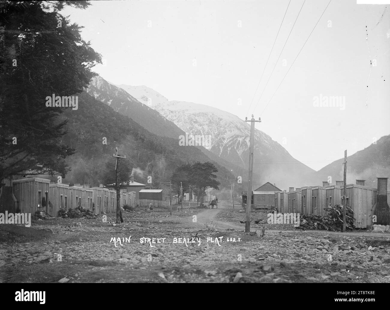 Vue de Bealey Flat, vue de Bealey Flat donnant sur main Street. Deux rangées de cabanes bordent la rue de chaque côté, avec des bâtiments plus grands au centre. Au-delà de Bealey Flat se trouvent des montagnes enneigées. P CA 1910 Banque D'Images