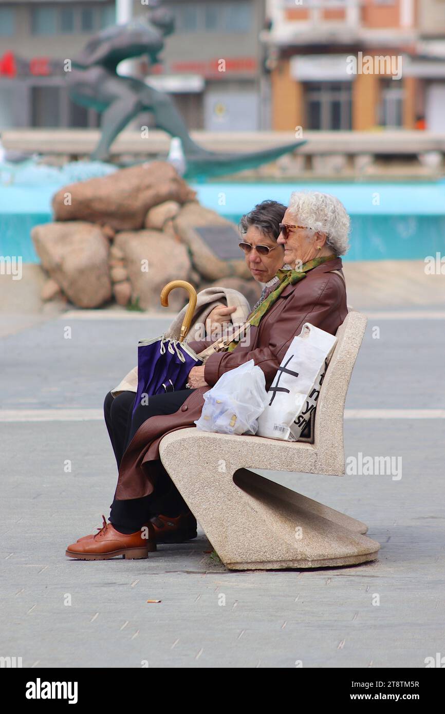 Deux dames âgées reposant en conversation sur un banc public à côté de la fontaine de Los Surfistas sur le hall de la plage de Orzán, la Corogne, Espagne. Banque D'Images