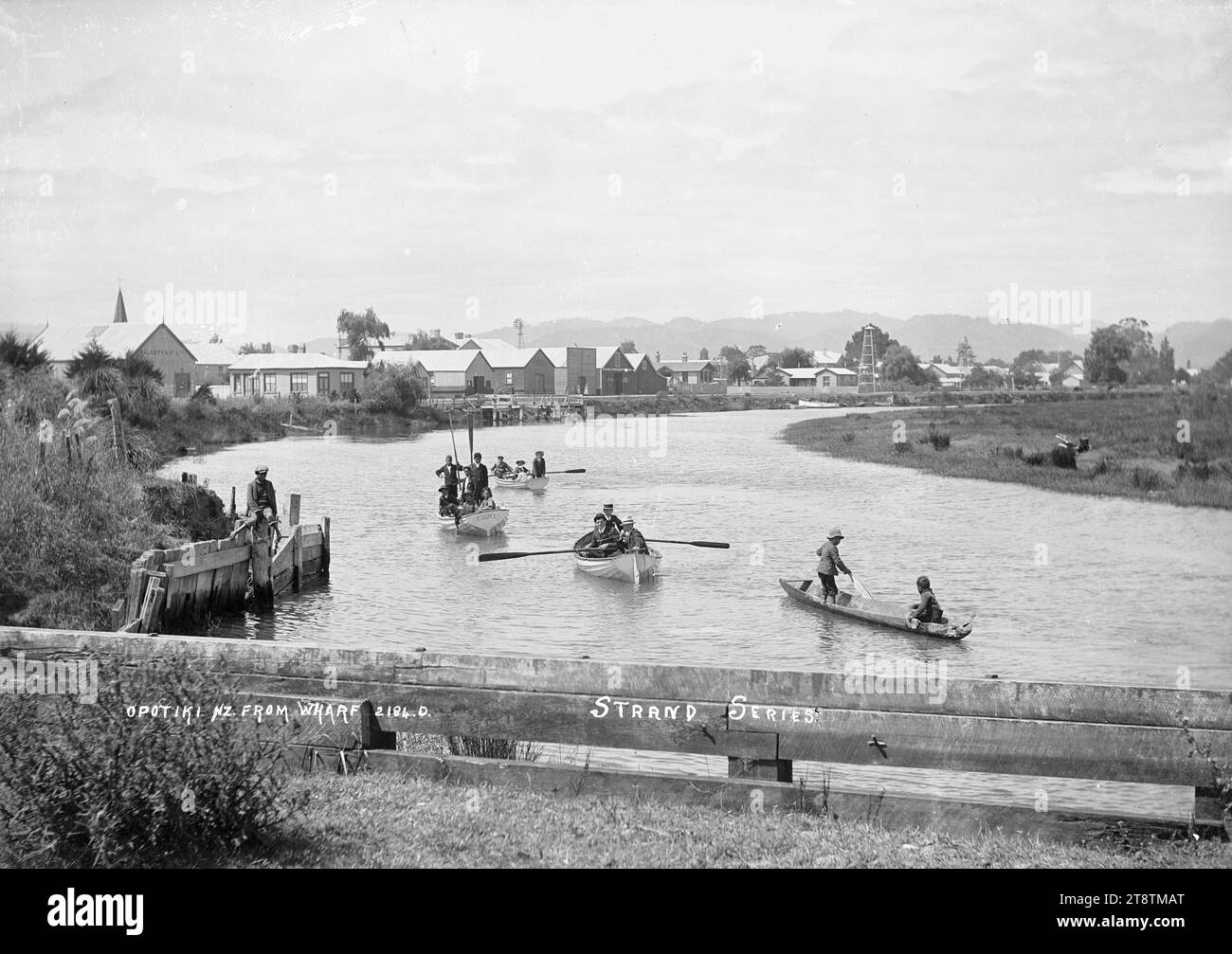 Vue d'Opotiki depuis le quai, vue de la rivière Waioeka vue vers le sud depuis le quai avec le canton d'Opotiki au loin. Trois chaloupes et un canot creusé sont au premier plan. Le bateau à rames avec le nom `Puhi' peint sur la poupe a six enfants maoris à bord, et deux des garçons sont debout et tiennent les rames debout. Un garçon maori se trouve sur la rive gauche au début des années 1900 Banque D'Images