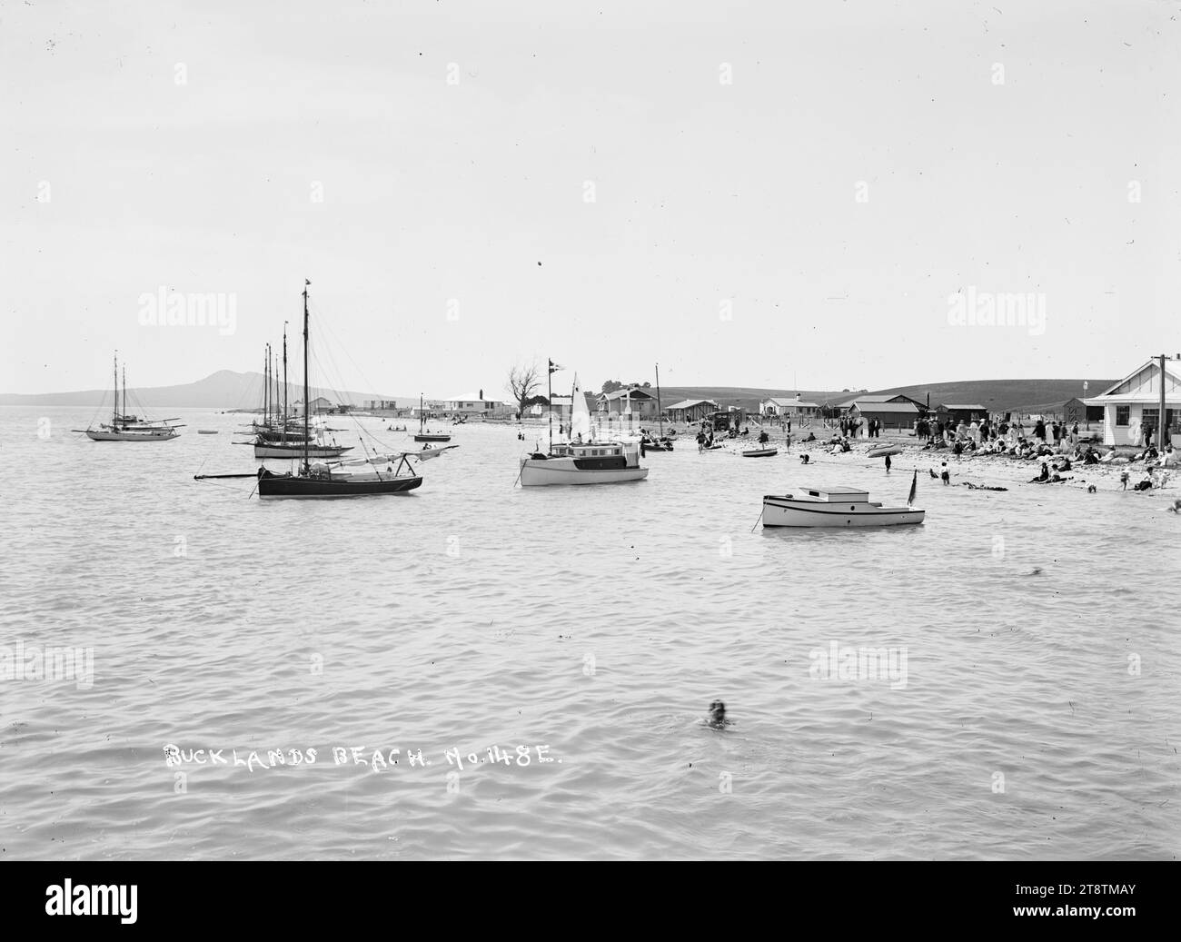 Artisanat de loisirs et vacanciers à Bucklands Beach, Auckland, Nouvelle-Zélande, vue de Bucklands Beach prise sur une journée d'été de la mer regardant vers la plage avec l'île de Rangitoto au loin. Les yachts et les lancements sont ancrés près du rivage et les gens sont sur la plage ou marchent le long du front de mer. Le magasin de Hindman est à l'extrême droite. Un homme nage près du photographe au premier plan au début des années 1900 Banque D'Images