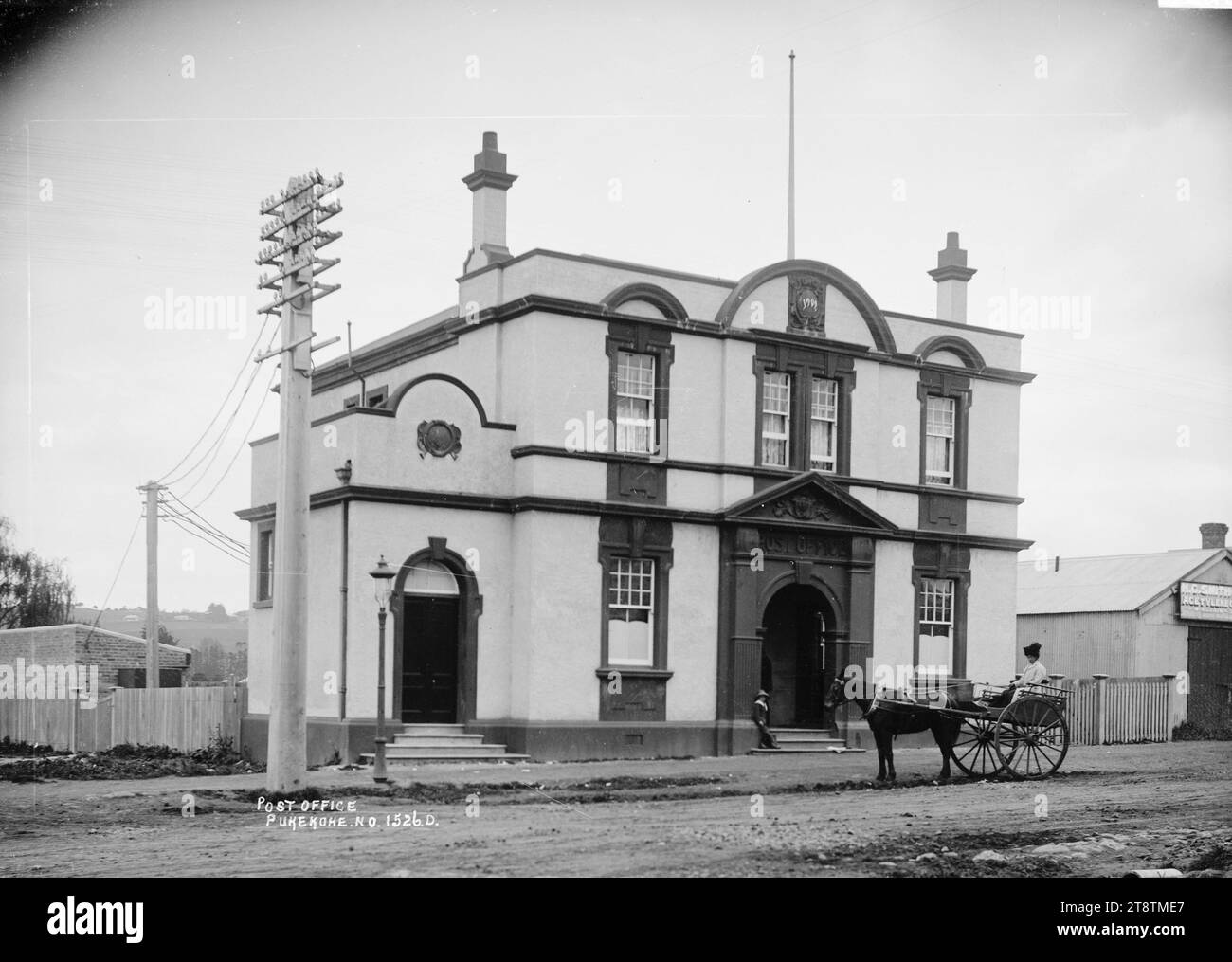 Bureau de poste de Pukekohe, vue du bureau de poste de Pukekohe dans la rue King prise après 1909. Un concert tiré par un cheval avec une femme est en face de l'entrée principale. À gauche, il y a une entreprise de plomberie. Un grand poteau télégraphique se trouve sur le côté gauche. Prise après 1909 Banque D'Images