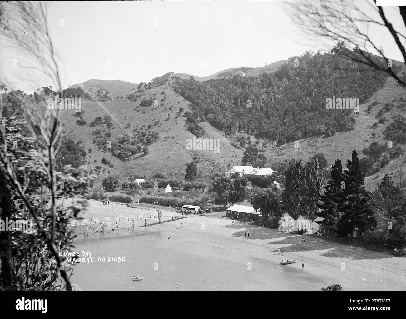 Hôtel et quai, Cowes Bay, Waiheke Island, vue de Cowes Bay vers le sud-ouest sur la plage depuis un point de vue élevé sur le promontoire au nord de la baie. Le quai peut être vu au premier plan central, et un grand bâtiment en bois (pension de Cowes Bay / hôtel?) est à mi-distance derrière les arbres. Les gens marchent sur la plage, rament ou jouent dans des bateaux près de la rive. Un groupe de gens achètent des choses à la boutique sur le front de mer en face du quai. Une tente est érigée dans le paddock devant la pension. Banque D'Images