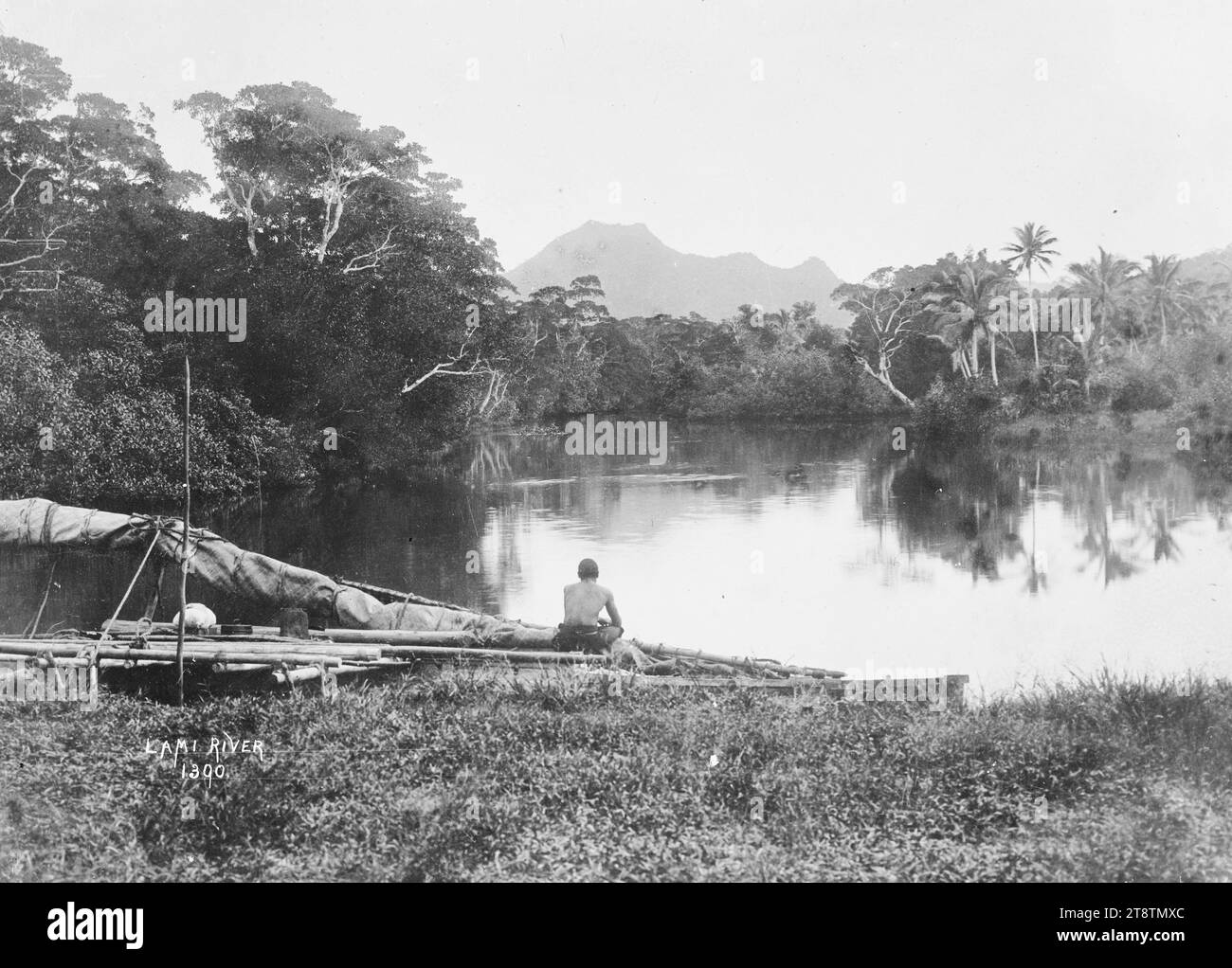 Canoë fidjien sur les rives de la rivière Lami, Viti Levu, Fidji, vue d'un canoë fidjien avec voile à fourrure au bord de l'eau sur la rivière Lami. Un homme est assis sur le canot et regarde la rivière. Les palmiers poussent sur la rive opposée et il y a des montagnes au loin. Prise au début des années 1900 Banque D'Images