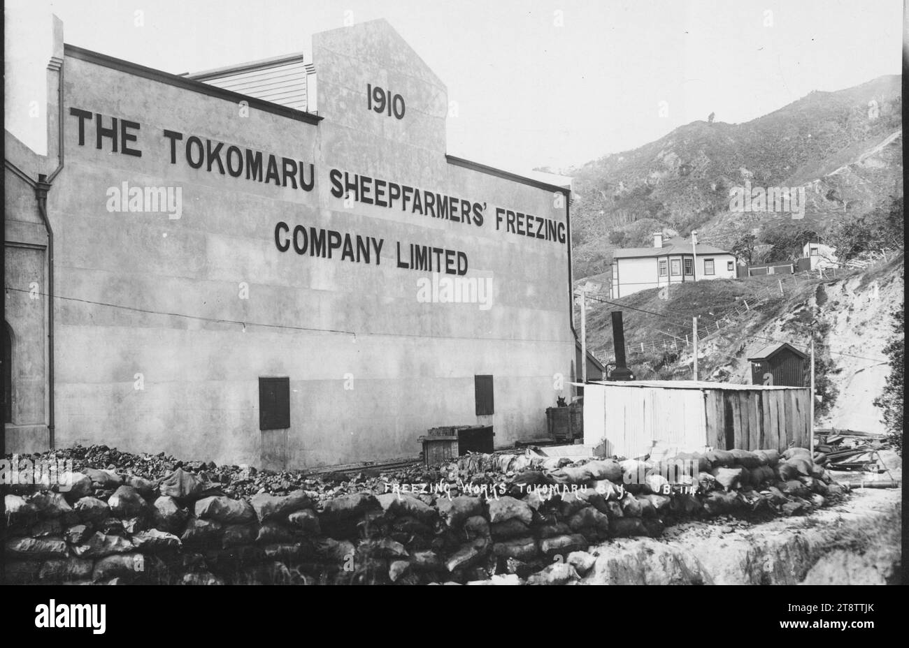 Tokomaru Sheepfarmers' Freezing Company Ltd travaille, à Waima, Tokomaru Bay, vue du bâtiment des travaux de congélation avec le nom et la date de son ouverture sur le devant. Les sacs sont empilés au premier plan. Sur la colline derrière se trouve la maison du directeur après 1910 Banque D'Images