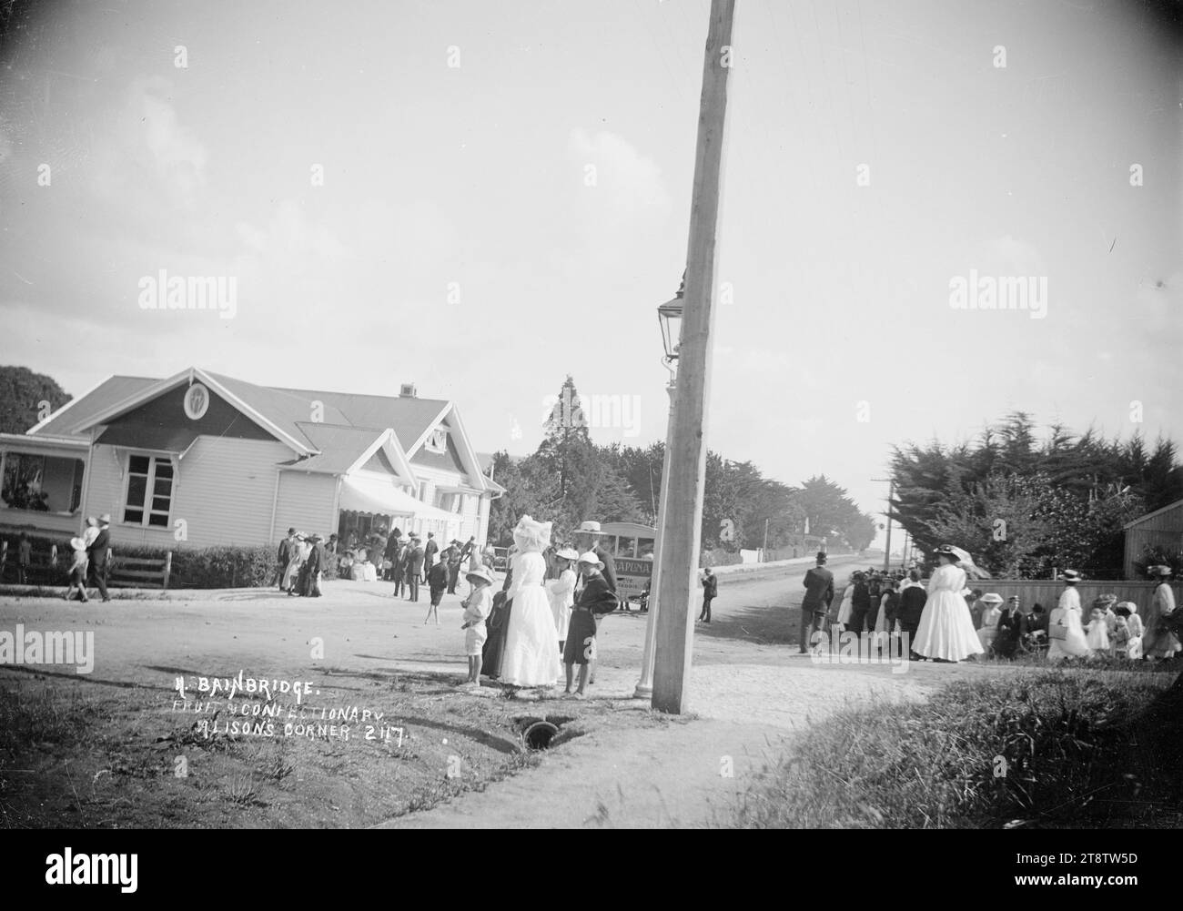 Bainbridge's fruit and Confectionery Shop, Alison's Corner, Takapuna, vue de la boutique de N Bainbridge sur Alison's Corner (maintenant connu sous le mon Desir Corner) avec des gens assis sous l'auvent de toile à l'avant de la boutique. Les gens se tiennent de l'autre côté de la route et un bus tiré par des chevaux attend à proximité. Le magasin semble faire partie d'une grande maison en bois de plain-pied. ca 1908 Banque D'Images