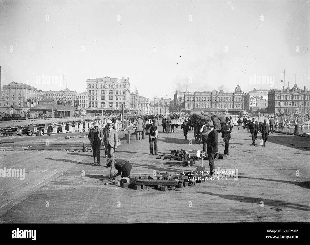 Queen Street Wharf, Auckland, Nouvelle-Zélande, prise au moment du remplacement de l'ancien Queen Street Wharf par un nouveau quai en béton ferro. Vue donnant directement sur Queen Street avec Endeans Building (construit en 1905) sur la gauche et le Gladstone Building (construit en 1883), avec Gladstone Coffee Palace, sur la droite. À l'extrême droite se trouve le siège social de l'Auckland, New Zealand Harbour Board ; tous dans Quay Street. Sur la gauche, de nouveaux pieux de béton sont enfoncés dans le lit du port. Au premier plan, les ouvriers remplacent les blocs de bois sur le vieux quai. Banque D'Images