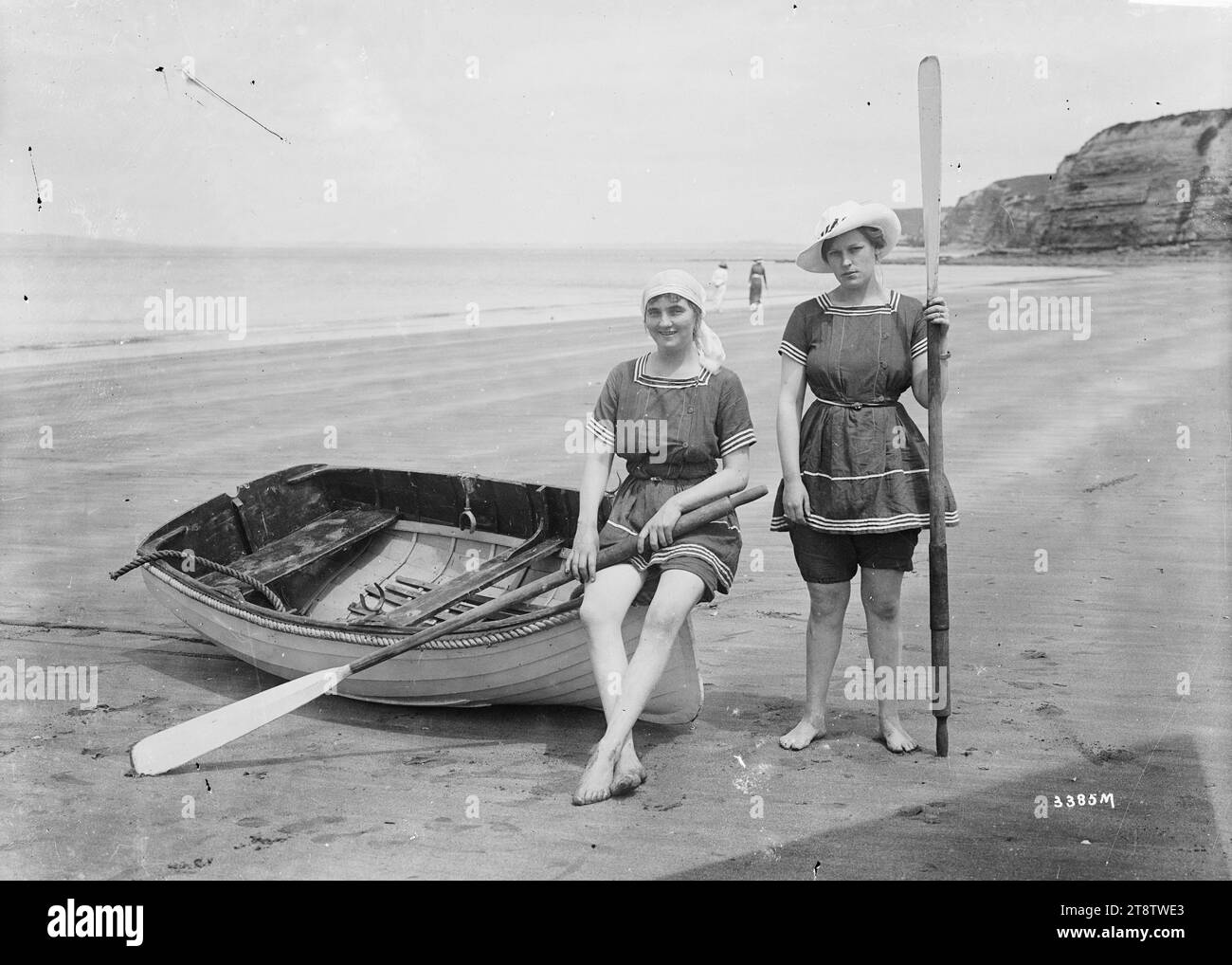 Deux femmes en costumes de bain, deux femmes sur la plage en costumes de bain. Les costumes sont tous deux composés d'une tunique à col carré, manches courtes, ceinture avec des rayures autour du cou et à la base des manches. Une paire de shorts sont portés en dessous. Une femme est assise sur une dighy tenant une rame. L'autre se tient à côté d'elle, tenant également une rame. Prise dans les années 1910 Banque D'Images