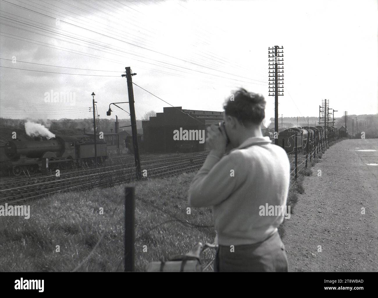 Années 1950, historique, un homme debout près d'une clôture prenant une photographie d'une locomotive à vapeur (no 62615) sortant d'une voie d'évitement de chemin de fer, Angleterre, Royaume-Uni. Banque D'Images