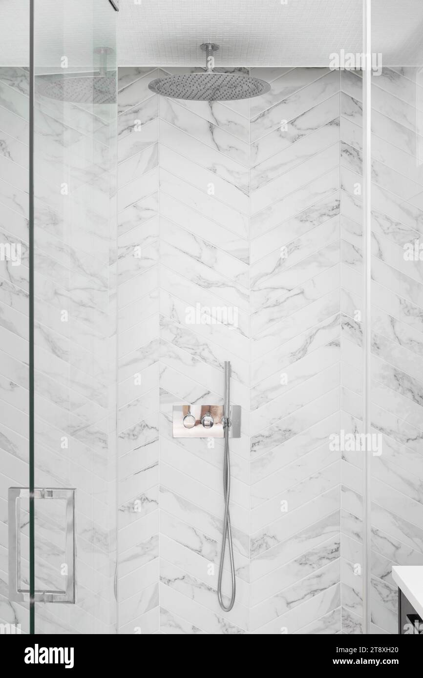 Un détail de douche de luxe avec des carreaux de marbre à chevrons, pomme de douche chromée polie et une porte vitrée. Banque D'Images