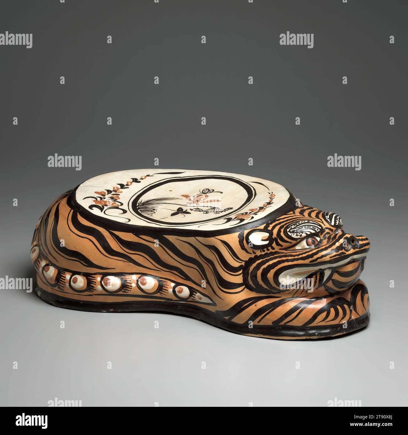 Coussin tigre, fin du 12e siècle, 4 7/8 x 14 7/8 x 7 po. (12,38 x 37,78 x 17,78 cm), TZ'u-chou ware grès avec glaçage noir et tan sur une barbotine blanche sous un glaçage clair, Chine, 12e siècle, cet oreiller amusant est modelé sous la forme d'un tigre accroupi, avec la tête retournée et les crocs dénudés. Le corps est recouvert de glaçure ambre-orange, qui, à son tour, est peint avec des bandes noires vives donnant un aspect naturaliste et une touche rythmique. Le haut de l'appuie-tête est peint avec un médaillon central encerclant un papillon planant au-dessus d'une orchidée et d'une feuille de lotus, le tout flanqué de sprays floraux Banque D'Images