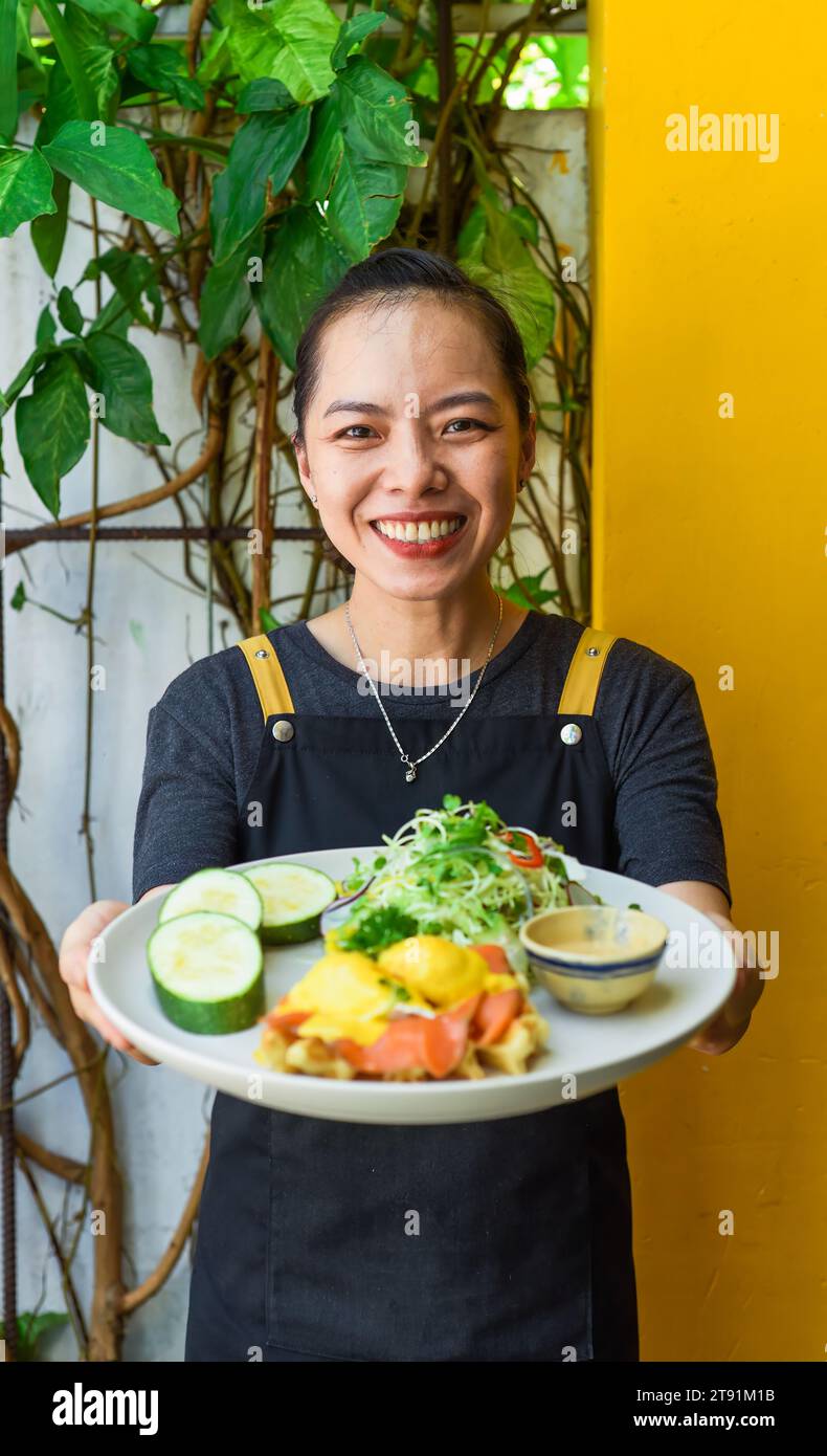 Jeune serveuse vietnamienne tenant une assiette avec des œufs bennedict gaufres avec du saumon fumé dans le restaurant Banque D'Images