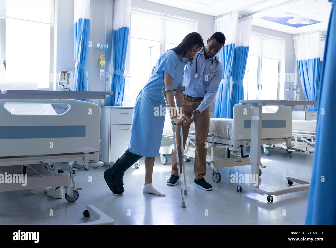 Patient féminin diversifié marchant avec des béquilles et médecin masculin aidant dans la chambre d'hôpital Banque D'Images