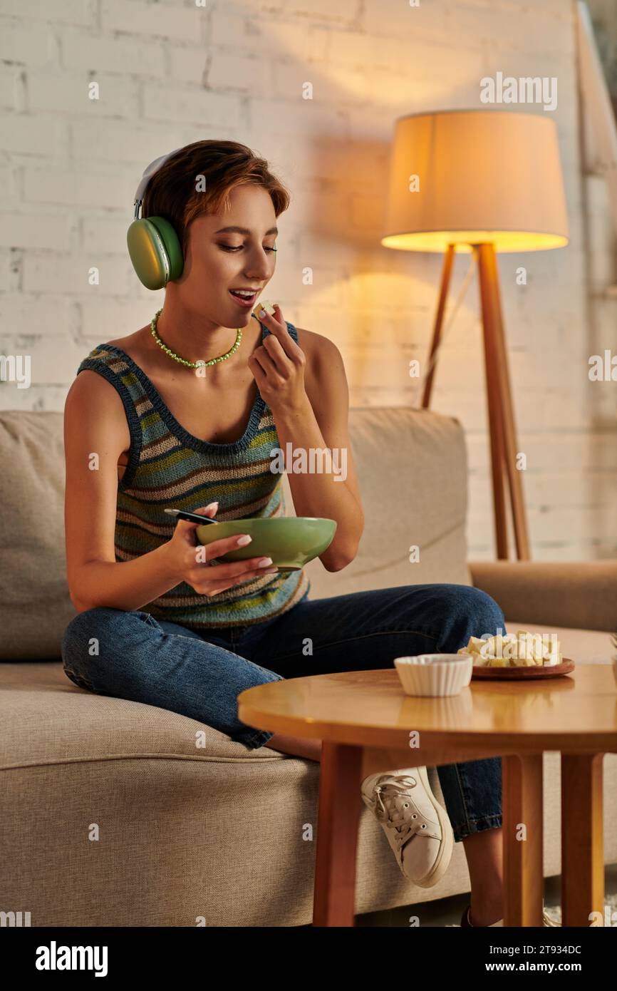 femme végétarienne dans les écouteurs tenant un saladier et mangeant du fromage tofu, collation du soir sur le canapé Banque D'Images