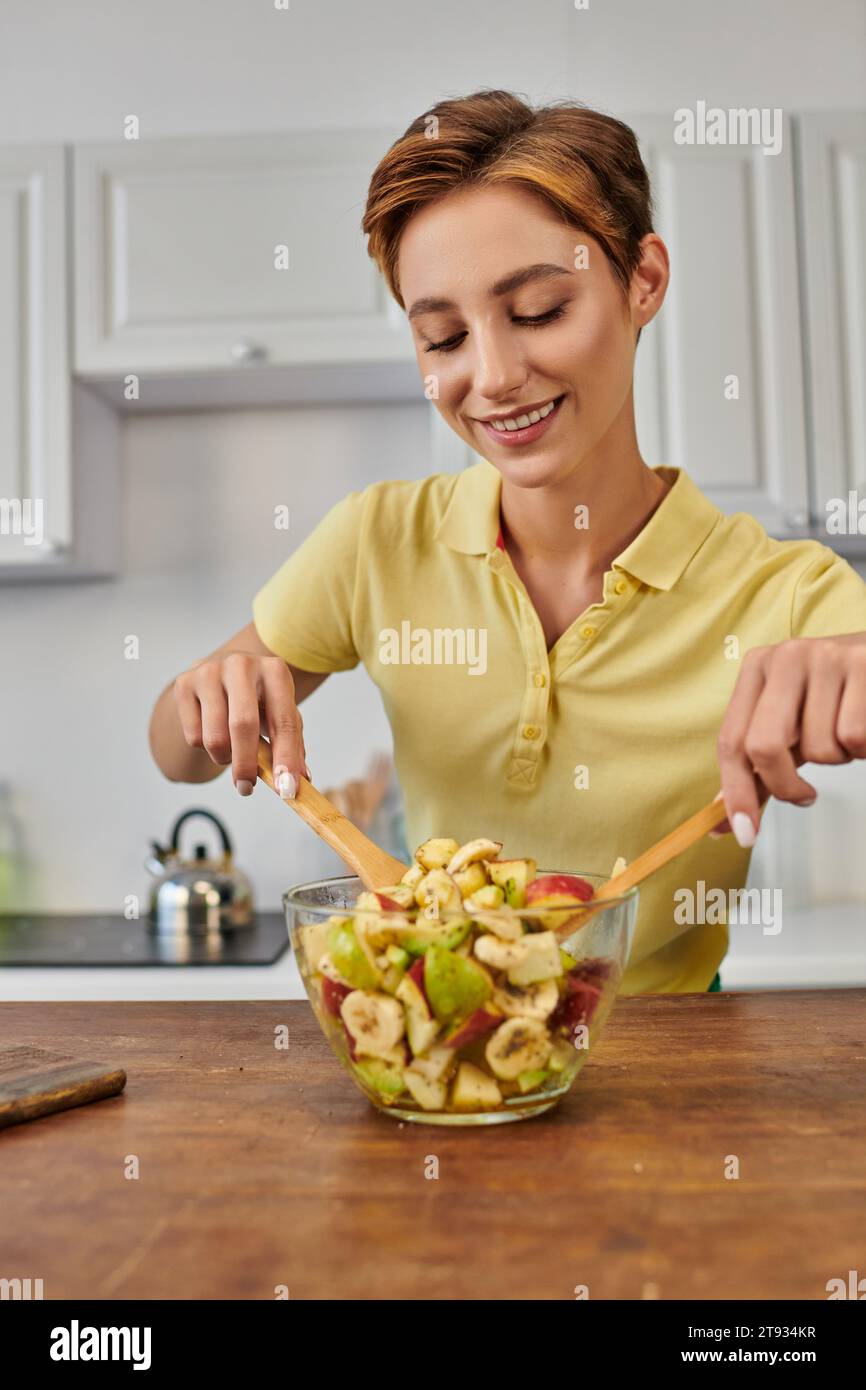 femme joyeuse mélangeant salade de fruits frais avec spatules en bois dans la cuisine, délicieuse recette végétarienne Banque D'Images