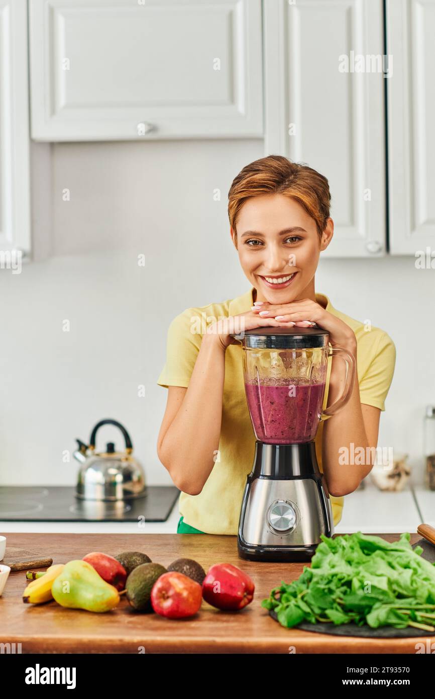 femme végétarienne souriant à la caméra près du mélangeur avec smoothie sain dans la cuisine, régime à base de plantes Banque D'Images