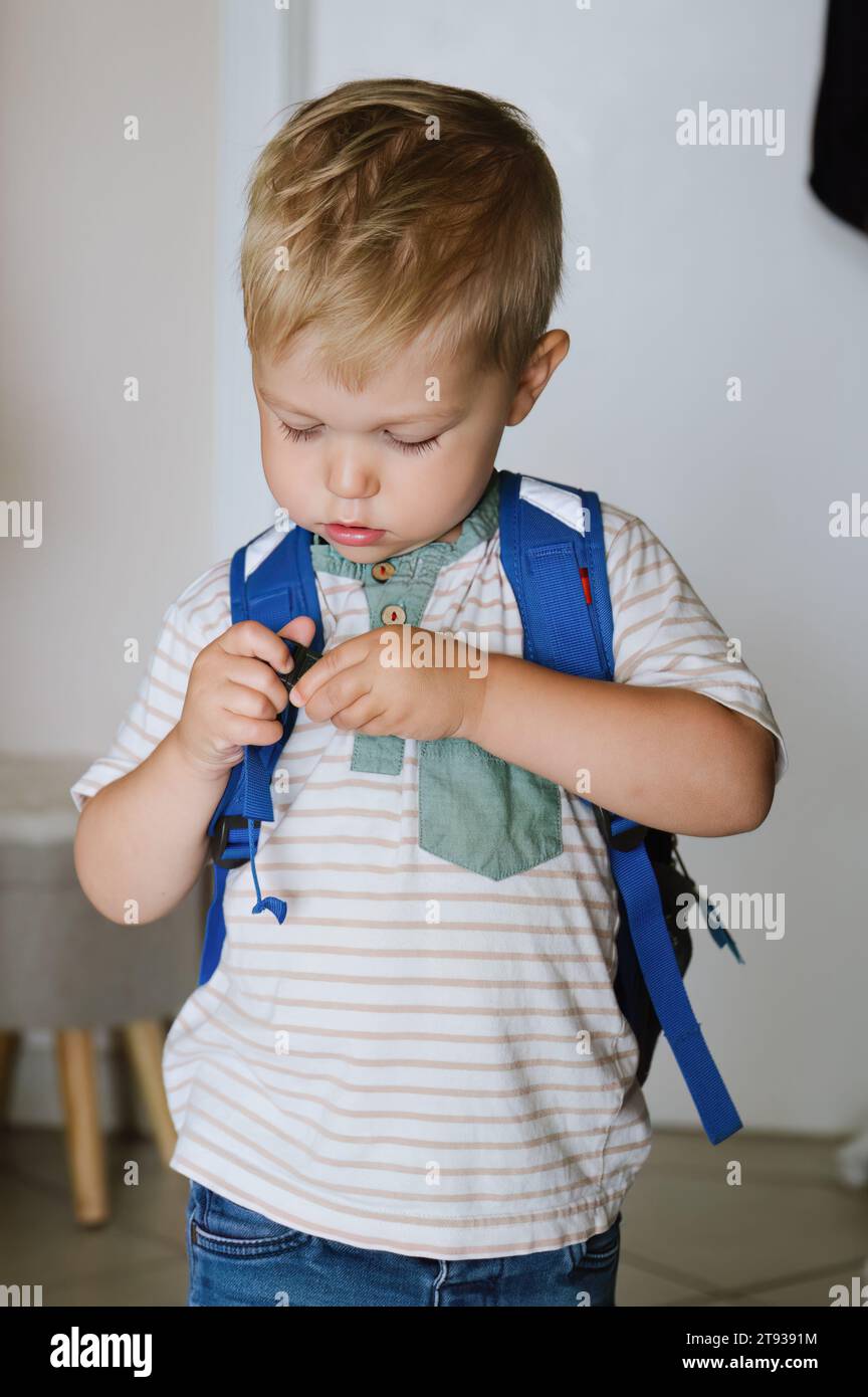 Adorable petit garçon dans des vêtements décontractés debout dans une pièce lumineuse et regardant vers le bas tout en ajustant le sac à dos Banque D'Images