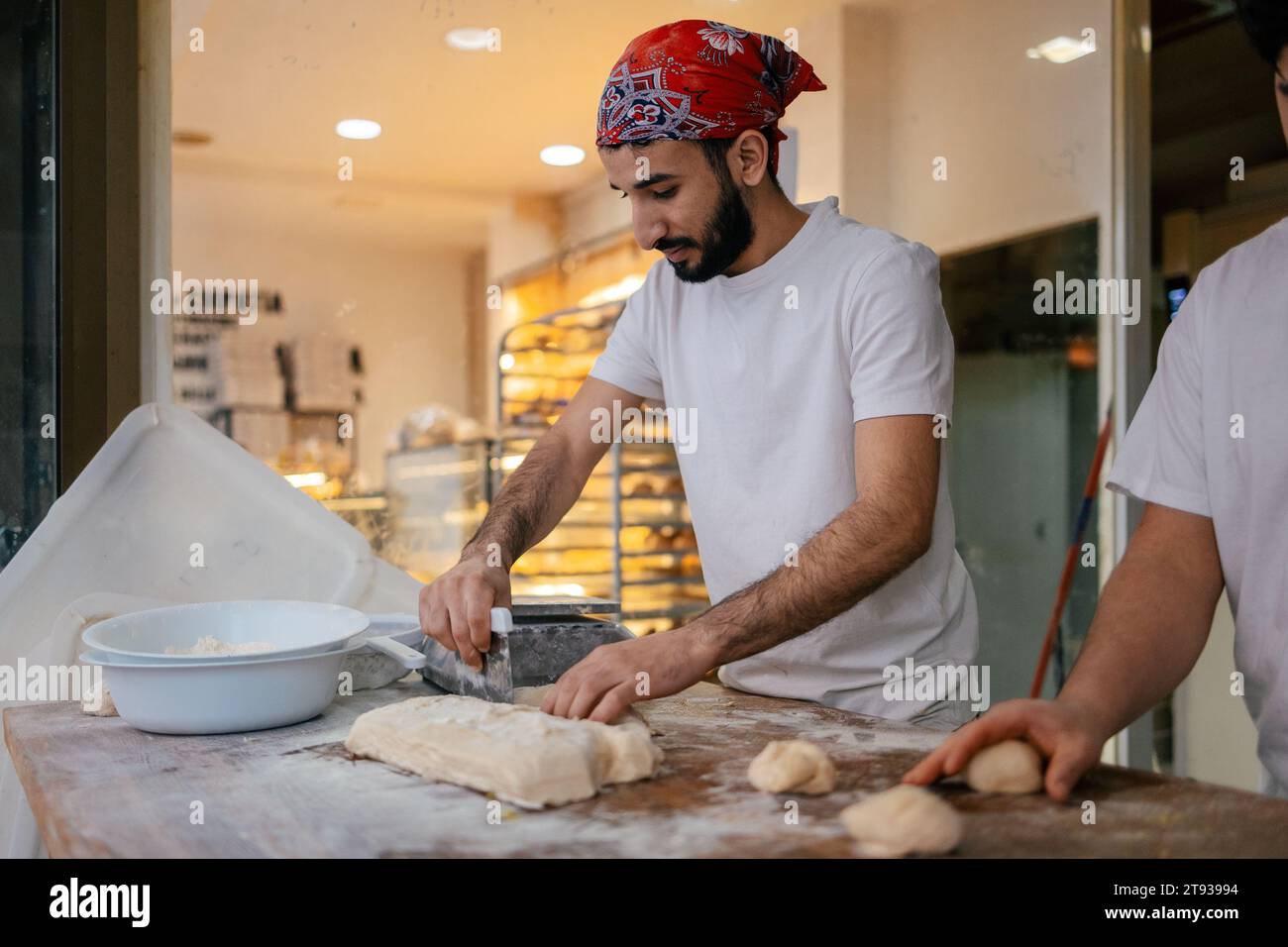 Jeune boulanger arabe habillé des vêtements blancs et un chapeau rouge coupant la pâte avec une omoplate dans une boulangerie Banque D'Images