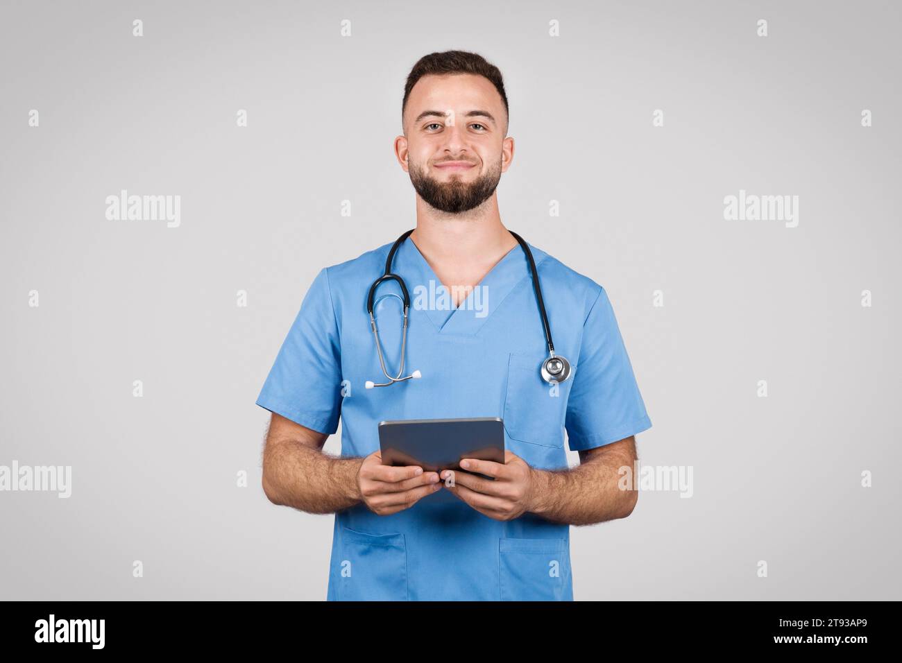 Médecin homme souriant avec comprimé, manteau bleu, fond gris Banque D'Images