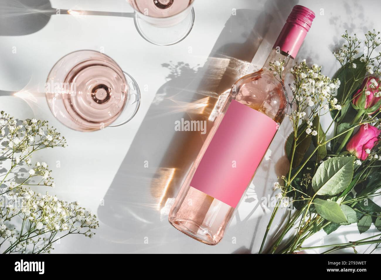 Bouteille de vin rose avec étiquette rose vierge, deux verres à vin et bouquet de fleurs romantique sur fond blanc avec lumière du soleil et ombre. Vin moderne Banque D'Images