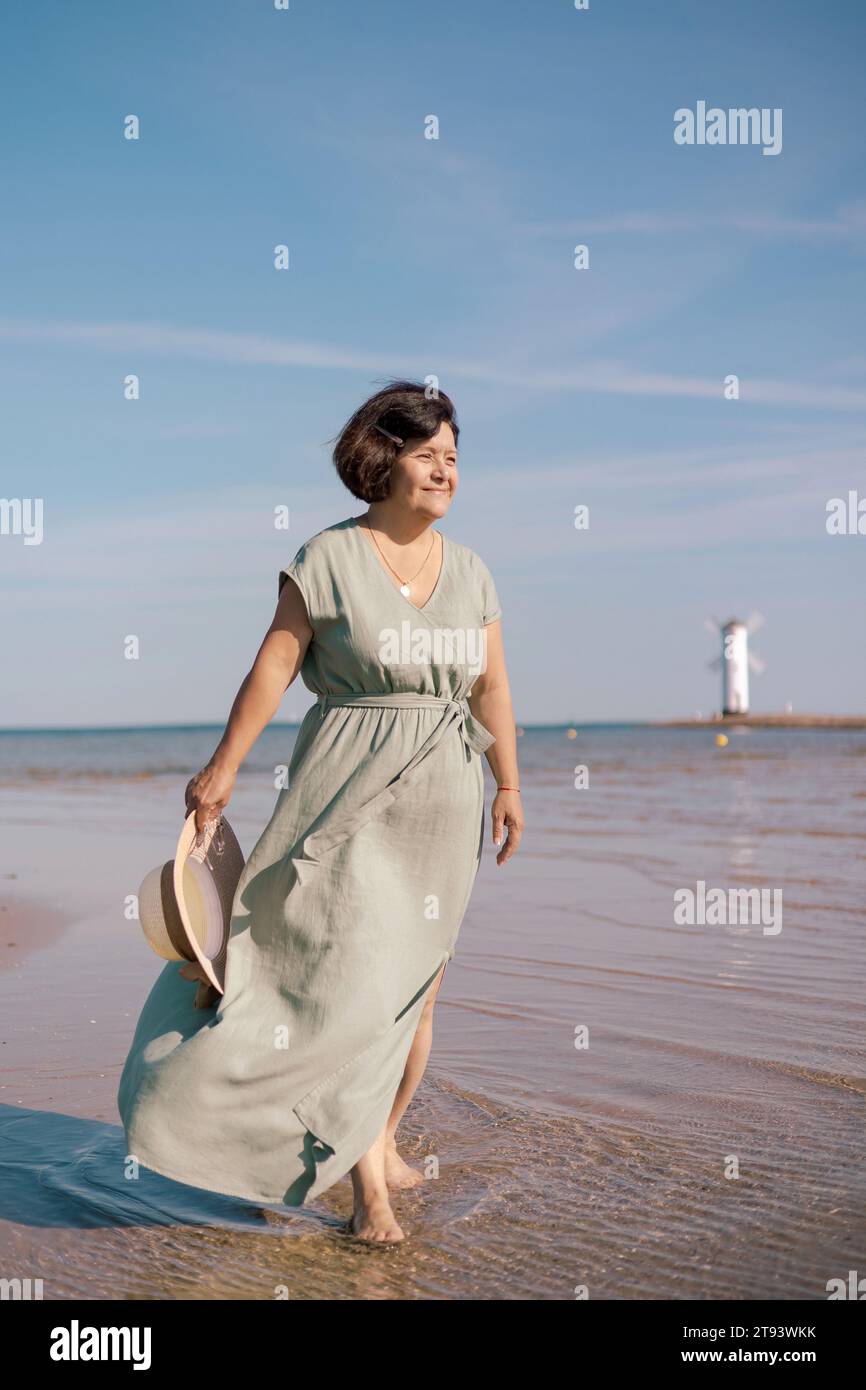 Une femme dans une robe fluide se tient sur une plage de sable du soleil avec un chapeau de soleil à la main Banque D'Images
