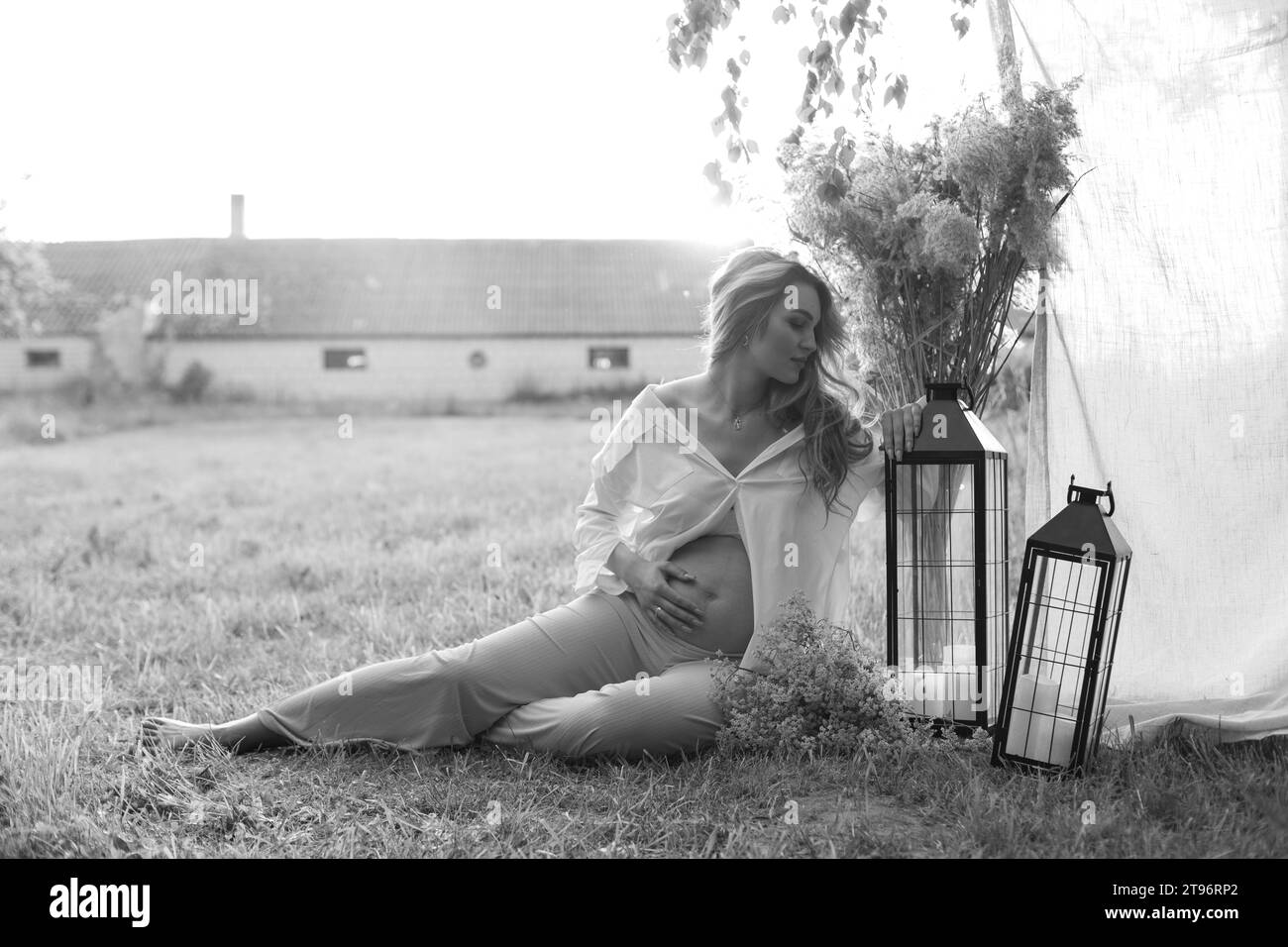 Portrait noir et blanc de belle femme enceinte assise sur l'herbe fauchée , elle embrasse doucement son ventre. Maternité. Banque D'Images