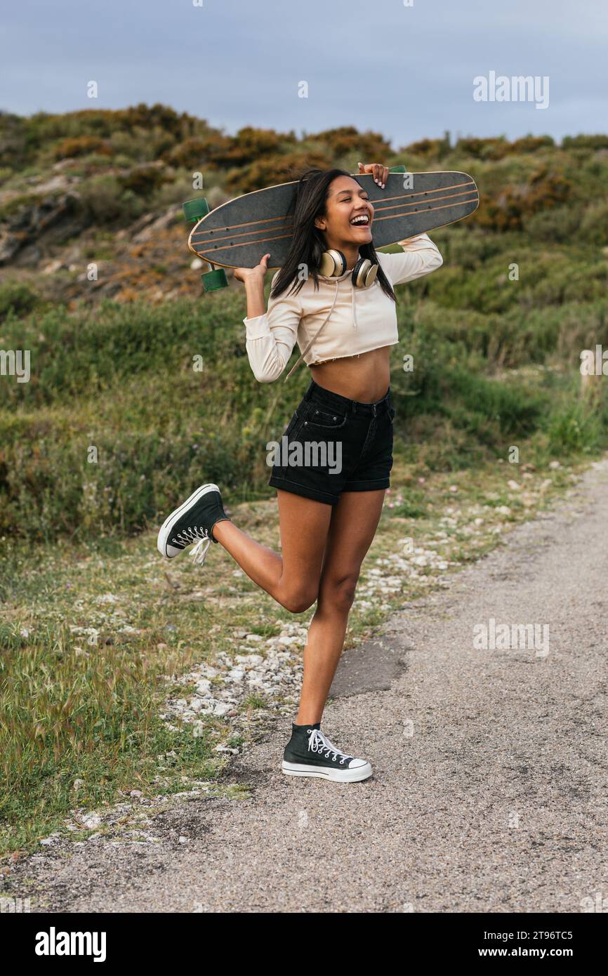 Corps complet de jeune femme gaie ethnique debout sur une jambe avec une longue planche derrière la tête et un casque sur le cou et rire avec la bouche ouverte Banque D'Images