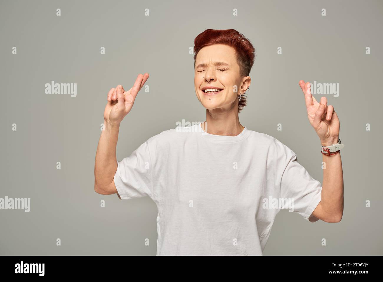 personne bigender tendue en t-shirt blanc croisant les doigts pour la chance debout avec les yeux fermés sur le gris Banque D'Images